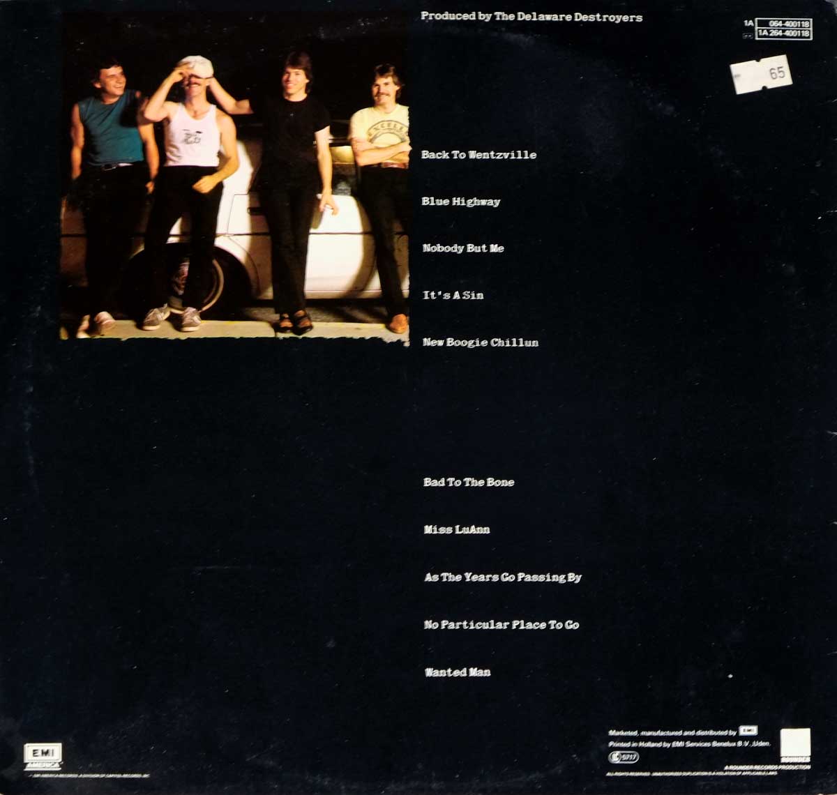 Back cover of the 1982 LP 'Bad to the Bone' by George Thorogood & The Destroyers. A candid photo of the four band members standing casually in front of a white car, with the tracklisting printed on a black background in distressed white text and a price sticker in the top right corner.