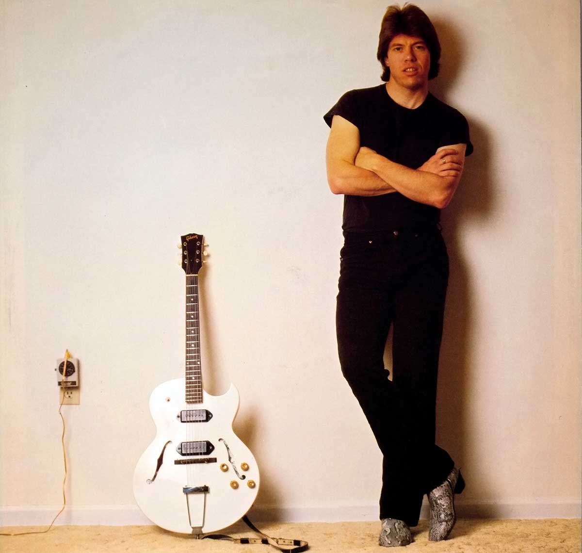 George Thorogood leaning against a plain wall with arms crossed, dressed in black, standing beside a white Gibson hollow-body electric guitar on the floor plugged into a nearby wall outlet, featured on the custom inner sleeve of the 1982 album 'Bad to the Bone.'