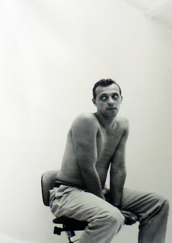 This is a black-and-white portrait of a Tom Novembre seated on a stool or chair, looking off to his left with a slightly contemplative or detached expression. The man, appears shirtless, with his arms resting on his lap and his shoulders slightly hunched forward, giving him a relaxed but introspective posture. The background is minimal and plain, allowing focus to remain on the subject. The photo has an intimate, almost vulnerable feel, enhanced by the simplicity of the setting and his unguarded expression. The lighting is soft, creating gentle shadows that emphasize his physique and facial features, capturing a mood that feels both introspective and quietly intense. https://vinyl-records.nl/