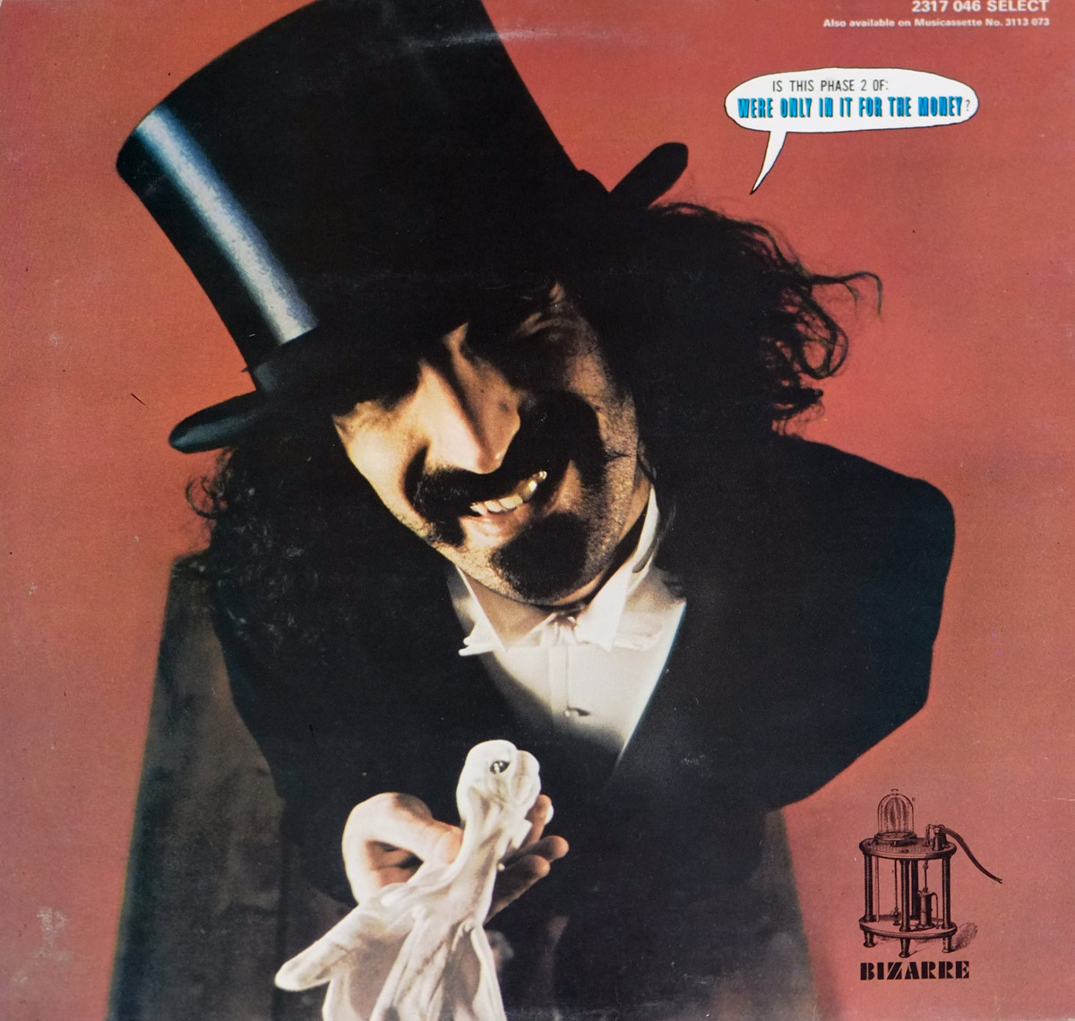 A dramatic portrait of Frank Zappa wearing a tall black top hat, leaning slightly forward with a wide grin and expressive eyes. The figure is dressed in a dark formal jacket with a white bow tie, set against a warm red background. The lighting creates deep shadows around the face and hat brim, adding theatrical impact. In the figure’s left hand, a white cloth or glove is extended outward as if being presented or offered. A small printed speech bubble near the upper right corner asks a cryptic question about a phase and money. The lower right shows a Bizarre Records illustration logo, featuring an old-fashioned mechanical contraption in a round frame. The overall composition blends humor, surrealism, and showman energy.