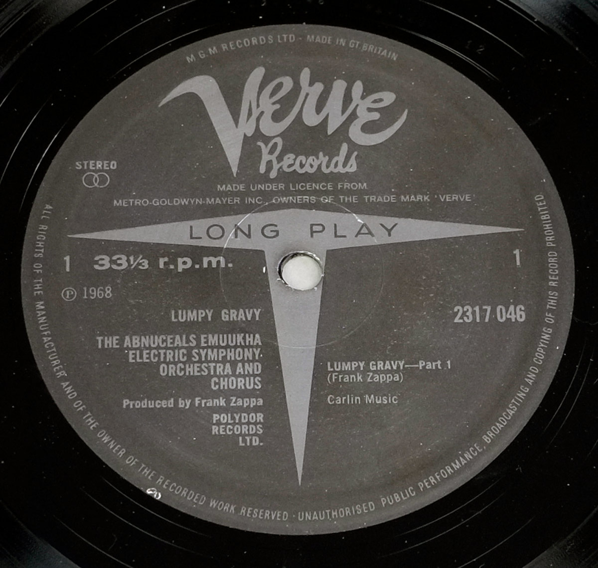 Close-up of the UK Verve Records Side One label for the album “Lumpy Gravy,” showing a dark grey background with the large Verve script logo at the top, a long-play arrow graphic through the spindle hole, catalog number 2317 046, 33⅓ r.p.m. speed marking, credits for The Abnuceals Emuukha Electric Symphony Orchestra and Chorus, and detailed rim text indicating manufacturing in Great Britain under licence from Metro-Goldwyn-Mayer.