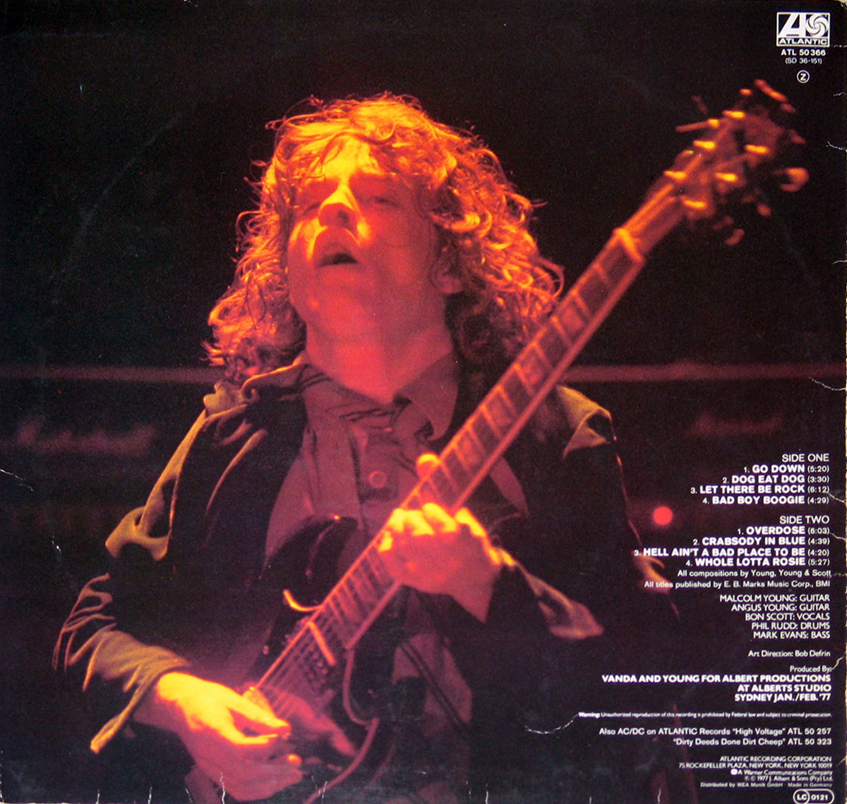 Back cover of AC/DC’s 'Let There Be Rock' (German Release) featuring a close-up performance shot of Angus Young bathed in fiery red stage light, mid-guitar solo, with curls flying and eyes half-closed in intensity. The image dominates the left side, while the right side lists track titles for both sides of the LP in white text, along with production credits, band member roles, and label logos for Atlantic Records. The visual captures the raw energy and passion of a live AC/DC performance.
