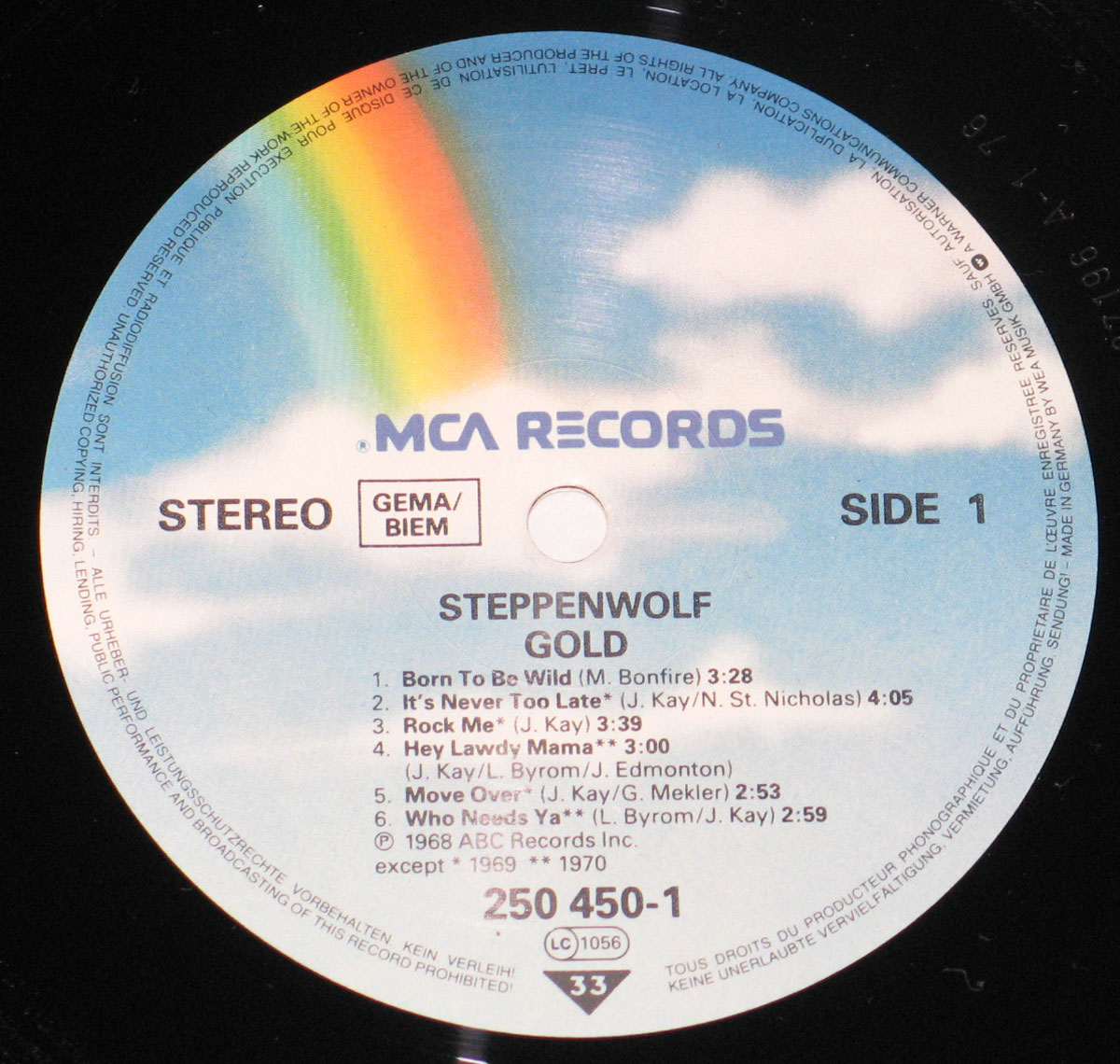 Close-up of the record label on Steppenwolf's Gold Their Great Hits (1970 European MCA Records release), Side One. The label background is a sky-blue gradient with soft white clouds and a vivid rainbow descending diagonally from the top left. The MCA Records logo is printed at the top in bold blue lettering. Below it, the text identifies the album title Gold and the band Steppenwolf. The tracklist for Side One is listed in black text, featuring six songs including Born To Be Wild, It's Never Too Late, Rock Me, Hey Lawdy Mama, Move Over, and Who Needs Ya, with composer credits and timings. The label also shows Stereo format, GEMA/BIEM rights society, catalog number 250 450-1, ℗ dates 1968 and 1969/1970 for tracks, and manufacturing details in Germany. The outer edge carries fine copyright warnings in multiple languages, completing the professional MCA rainbow-label style of the early 1970s.