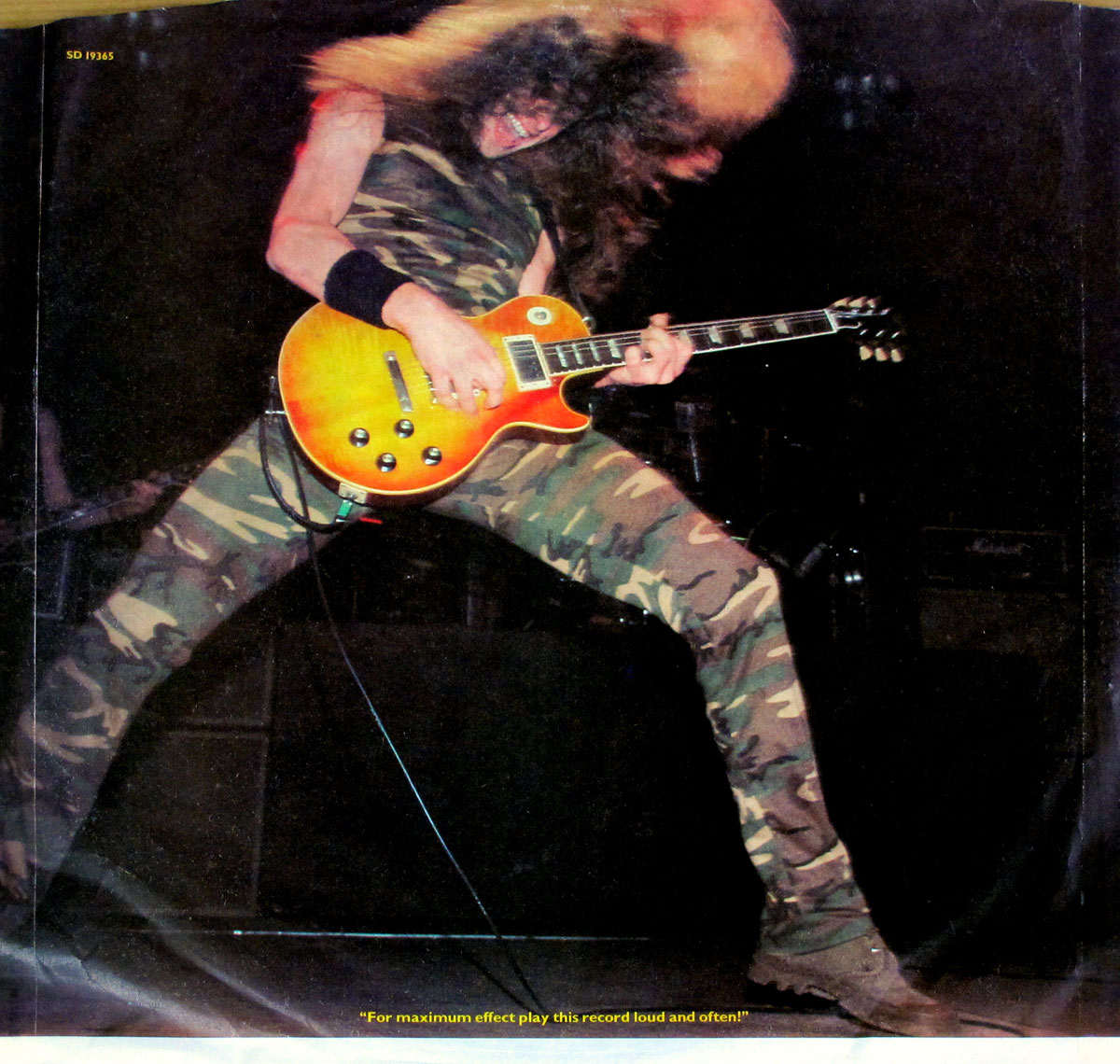 Reverse side of the custom inner sleeve for the 1982 Ted Nugent album Nugent, Atlantic ATL 50 898, showing a dynamic live action photo of Nugent in mid-performance. He is wearing a sleeveless camouflage jumpsuit, legs spread wide in a power stance, head thrown back with hair flying, and playing a sunburst Gibson Les Paul guitar under stage lights. The background is dark with faint outlines of amplifiers, and a caption at the bottom reads 'For maximum effect play this record loud and often!'