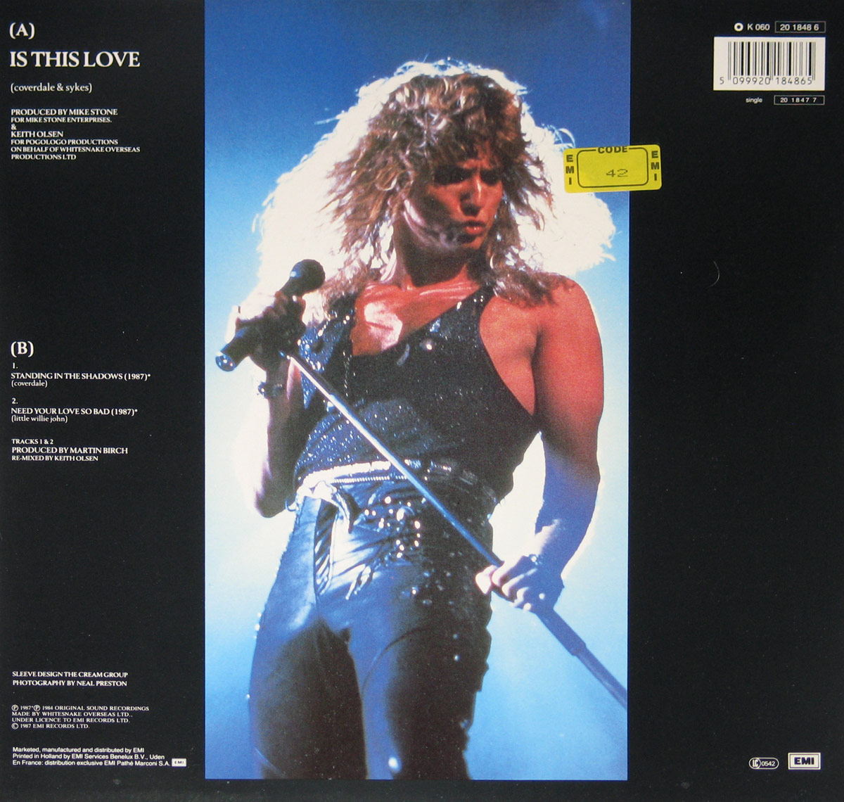 Back cover of Whitesnake’s 12-inch Maxi-Single 'Is This Love', featuring a dramatic live photo of David Coverdale mid-performance under intense blue stage lighting. The left panel lists track information for both sides of the record, production credits, and design acknowledgments. The overall layout contrasts dark negative space with the luminous, kinetic energy of Coverdale’s pose, his hand gripping the microphone stand as his hair catches the backlight, symbolizing the raw charisma that defined Whitesnake’s late-1980s aesthetic.