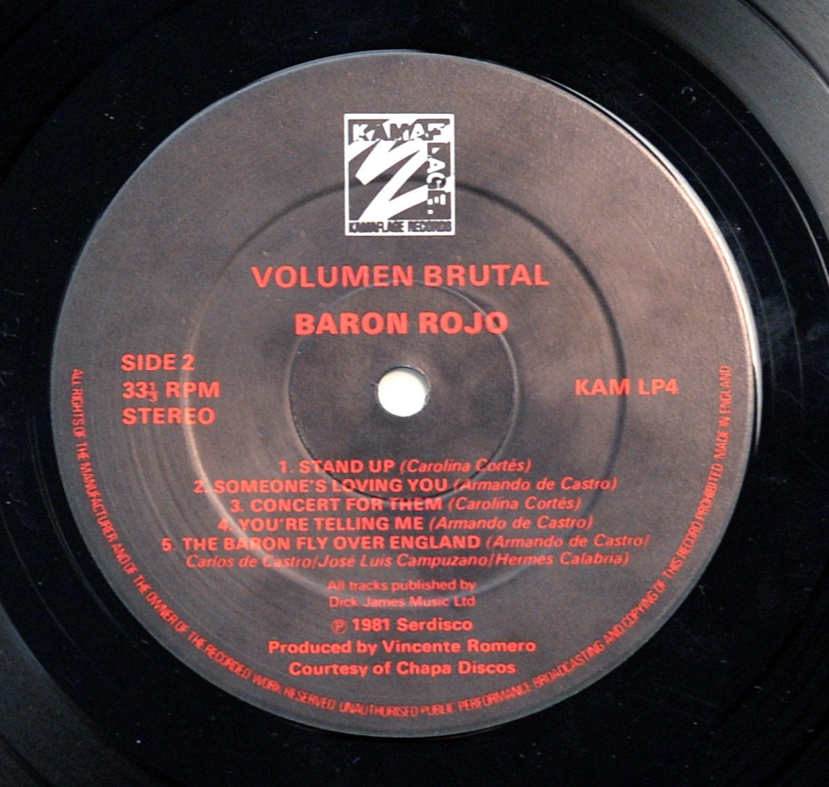 High-resolution close-up of Side Two record label for Barón Rojo’s 1982 album Volumen Brutal, issued by Kamaflage Records in the UK. The matte black label features red text and the Kamaflage logo printed at the top center. Below the logo, the album title Volumen Brutal and band name Baron Rojo are printed in bold uppercase letters. On the left, SIDE 2, 33⅓ RPM, and STEREO are neatly aligned, while the catalog number KAM LP4 appears on the right. The track listing includes five songs: Stand Up, Someone’s Loving You, Concert for Them, You’re Telling Me, and The Baron Fly Over England, with songwriting credits attributed to Carolina Cortés, Armando de Castro, and band members José Luis Campuzano and Hermes Calabria. The lower section notes publishing by Dick James Music Ltd and production by Vicente Romero, courtesy of Chapa Discos. Around the edge, fine red text reads 'ALL RIGHTS OF THE MANUFACTURER... MADE IN ENGLAND,' typical of early 1980s heavy metal vinyl pressings.