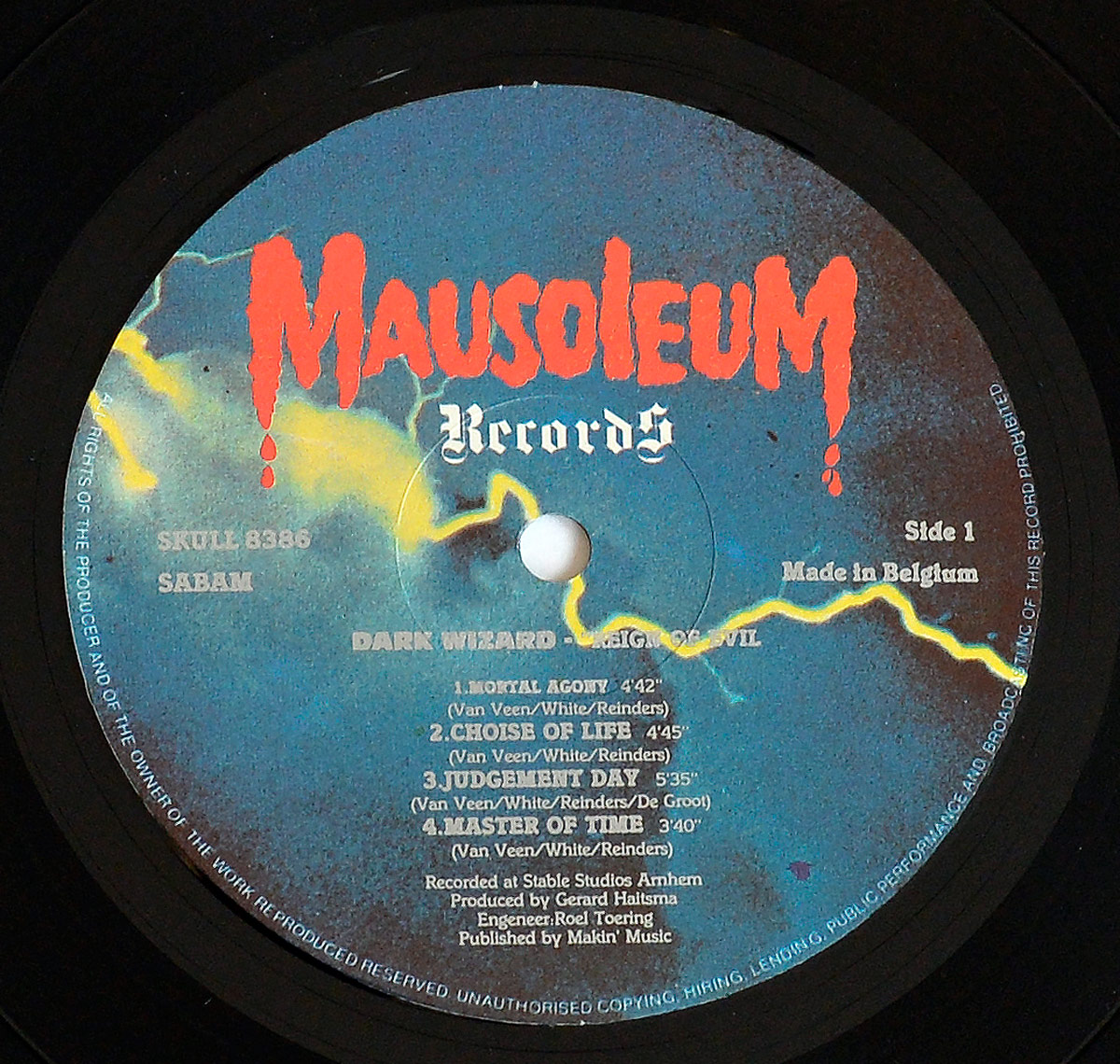 Close-up of Side One record label for Dark Wizard's 1985 album Reign of Evil on Mausoleum Records SKULL 8386. The label features a dramatic design with a stormy blue background and bold red Mausoleum Records logo dripping like blood. Track titles Mortal Agony, Choice of Life, Judgement Day, and Master of Time are listed with durations and songwriting credits. The label also includes SABAM rights info, 'Made in Belgium', catalog number, and credits for producer Gerard Haitsma and engineer Roel Toering.