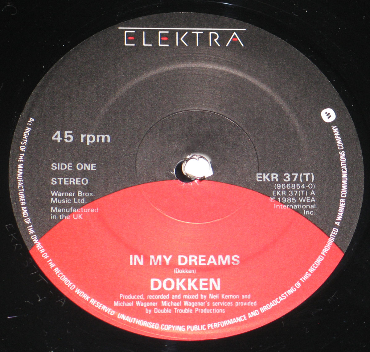 Close-up of the Elektra record label for Dokken’s 1985 UK 12-inch EP In My Dreams / Til The Livin’ End - Alone Again. The label design features Elektra’s black background with a bold red semicircle cutting across the lower half. At the top, the Elektra logo appears in white and red. Text on the left details Side One, stereo format, and Warner Bros. Music Ltd. manufacturing information. On the right, catalog number EKR 37(T) and publishing rights are printed. Centered within the red half, the track title In My Dreams and band name Dokken appear in white uppercase. Production credits list Neil Kernon and Michael Wagener.