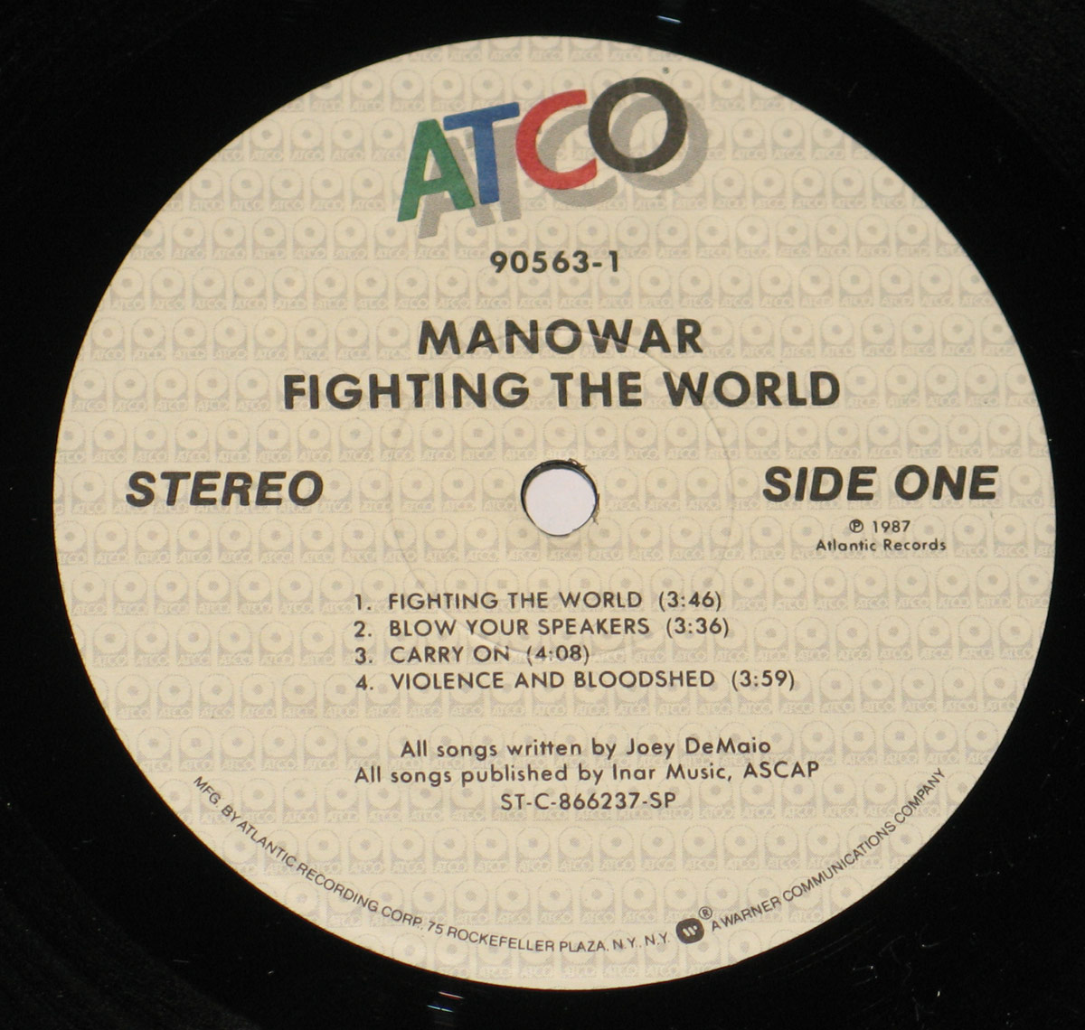 Close-up of the ATCO Records label for the 1987 USA vinyl pressing of Manowar's Fighting the World. The beige background is covered with a repeating ATCO logo watermark. At the top, the bold multicolor ATCO logo appears in green, red, blue, and grey. Below it, the catalog number 90563-1 is printed, followed by the album and band name in uppercase: MANOWAR FIGHTING THE WORLD. The text indicates Side One, Stereo, and copyright 1987 Atlantic Records. Four track titles are listed: Fighting the World (3:46), Blow Your Speakers (3:36), Carry On (4:08), and Violence and Bloodshed (3:59). At the bottom, credits state all songs written by Joey DeMaio and published by Inar Music ASCAP. Around the rim, the manufacturer information reads Manufactured by Atlantic Recording Corp, 75 Rockefeller Plaza, N.Y., N.Y., with Warner Communications Company logo.