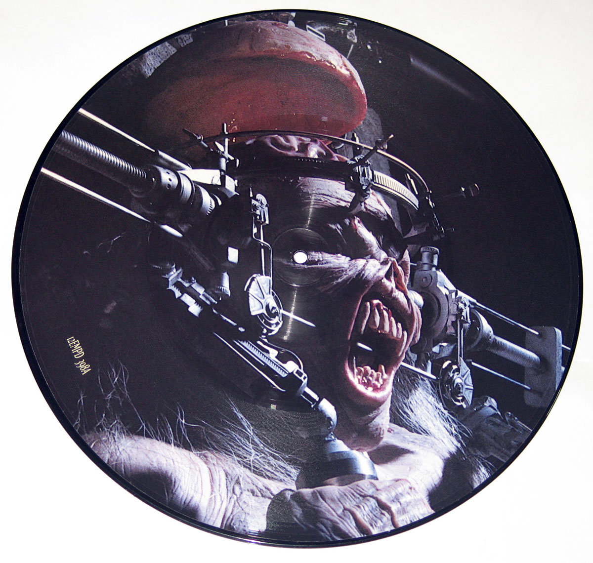 Close-up of a 12-inch picture disc: Iron Maiden’s mascot Eddie is shown in three-quarter profile, restrained in a mechanical head brace. The printed artwork spans the grooved vinyl; the center hole sits near the face. Eddie’s mouth is wide open in a scream, with exposed gums and jagged teeth, while steel arms, clamps, and bolts frame his head. A reddish cranial section appears like an opened cap above the brace. Lighting is cold and directional against a near-black background. Tiny catalog text is printed near the rim.