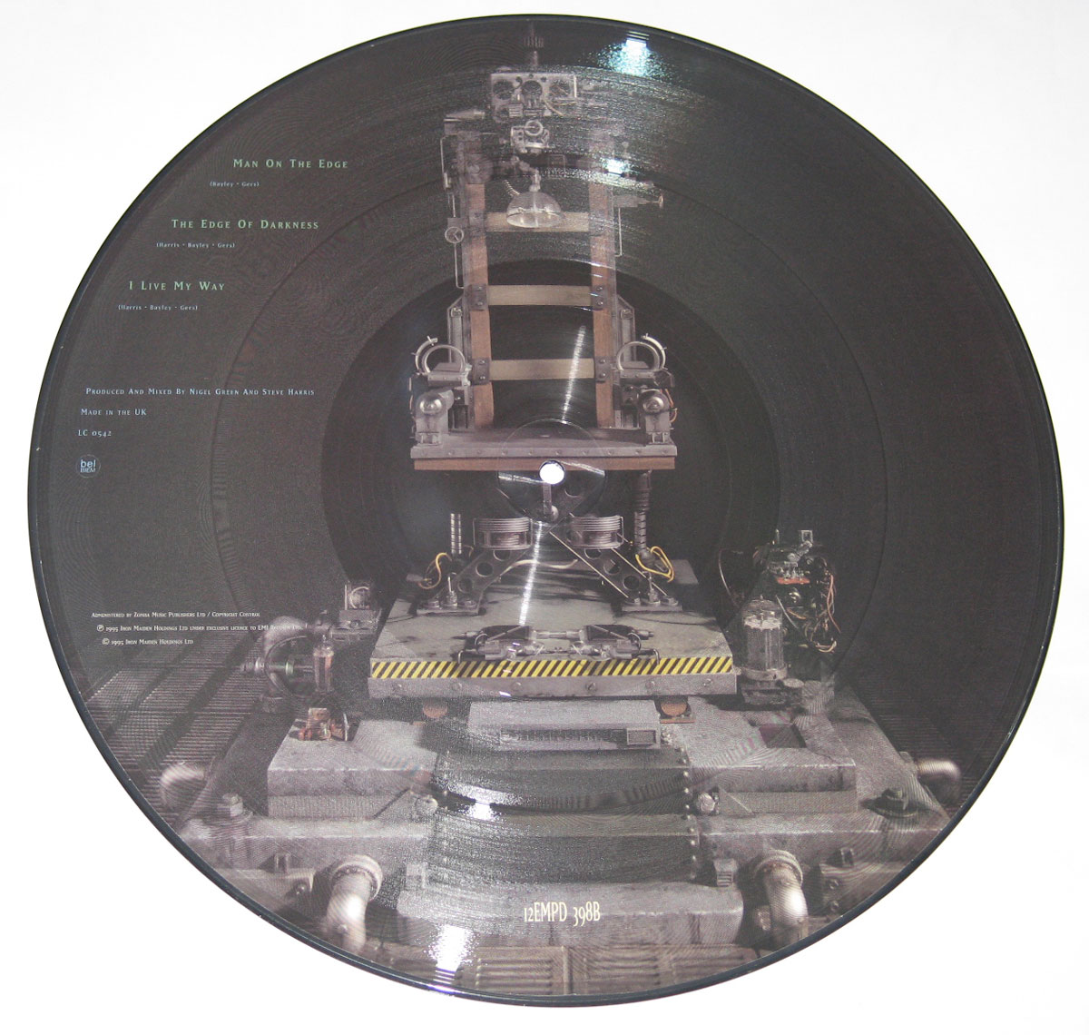 12-inch picture disc, side two. Printed artwork shows a front-facing industrial restraint chair set on a tracked platform inside a tunnel-like bay. The vinyl’s spiral grooves radiate around the scene. At left, a tidy column of small text lists tracks—Man On The Edge, The Edge Of Darkness, I Live My Way—followed by brief production credits and Made in the UK. The center hole aligns with the chair’s seat mechanism. Hazard stripes, pistons, bolts, and cabling detail the machine base; thin catalog text appears near the lower rim. Palette is grey, black, and steel.