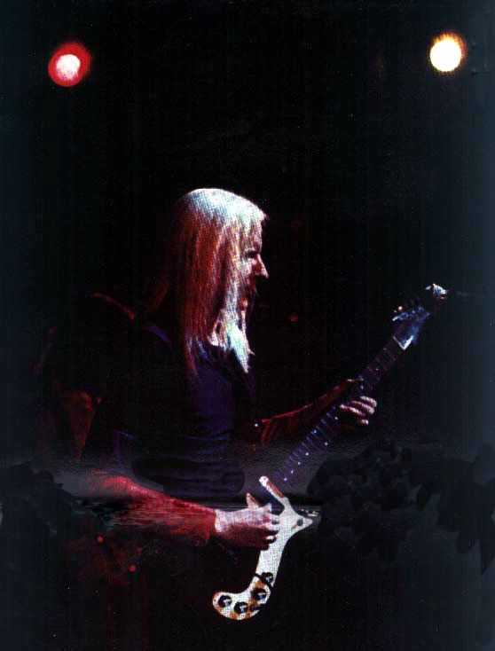 Johnny Winter in Paris (France), the legendary blues-rock guitarist, performing on stage. He has long, straight, white-blond hair, and his posture is focused as he plays. The setting appears dark with two stage lights above, casting a warm and moody glow over him.