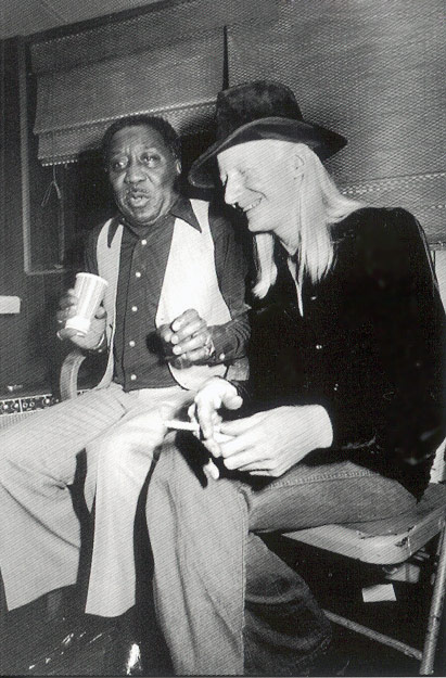 Black-and-white photo of blues legends Muddy Waters and Johnny Winter seated together, smiling and sharing a lighthearted moment. Muddy Waters holds a cup and wears a vest over a collared shirt, while Johnny Winter, with long hair and a broad-brimmed hat, sits beside him, appearing engaged in conversation. The casual setting suggests a backstage area, capturing their camaraderie and shared love for blues music.