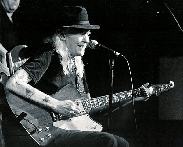 Black-and-white photograph of Johnny Winter seated on stage, playing an electric guitar. He wears a wide-brimmed dark hat and a short-sleeved shirt, with his long, light hair falling over his shoulders. His tattooed arms are clearly visible as he strums his guitar, a distinctive Gibson Firebird with a reverse headstock. Winter leans toward a microphone positioned in front of him, singing or speaking into it. Another musician can be faintly seen in the background playing bass. The setting suggests an intimate live performance, with the spotlight focused on Johnny Winter, highlighting his expressive posture and intense engagement with the music.
