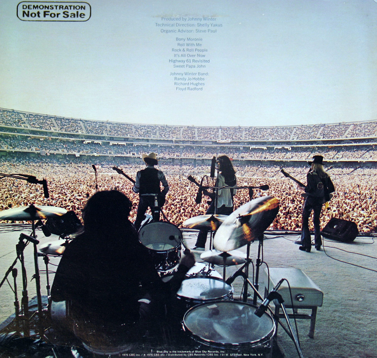 Back cover of the Promotional Copy of 'Captured Live' by Johnny Winter. The image captures a breathtaking perspective from behind the drummer on stage, looking out at a massive crowd packed into a stadium. The band is silhouetted against the bright sky as they play to an ocean of fans. Johnny Winter stands on the right with his signature long white hair, wearing a cowboy hat and playing his guitar. The band lineup is visible at center stage, with a microphone setup and multiple guitars. The top left corner of the cover bears the text 'DEMONSTRATION Not For Sale,' emphasizing that this is a promotional copy. Above the stage, the album's production and technical credits, along with the tracklist, are printed in a simple, understated font. The sheer scale of the audience and the dramatic stage view make this back cover an iconic representation of Johnny Winter’s electrifying live performances.