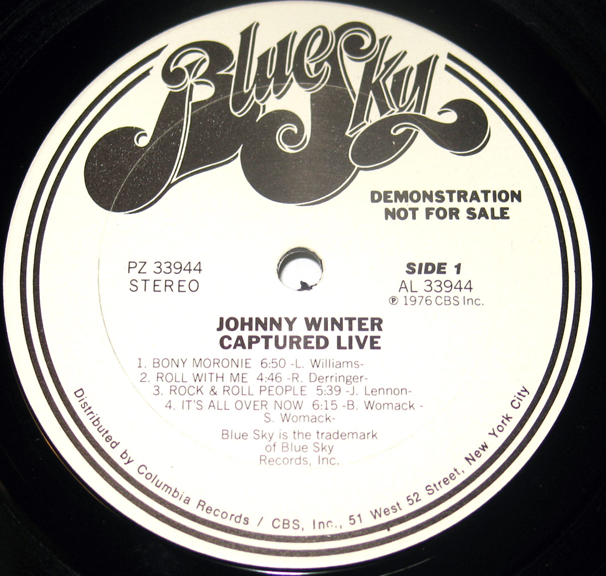 Close-up of Side One record label from the promotional copy of 'Captured Live' by Johnny Winter. The label is white with black lettering and prominently displays the 'Blue Sky Records' logo in stylized cursive at the top. The text 'DEMONSTRATION NOT FOR SALE' is printed in bold uppercase letters on the right side. Below, the album title 'Johnny Winter - Captured Live' is printed, followed by the tracklist for Side One: 'Bony Moronie' (6:50), 'Roll With Me' (4:46), 'Rock & Roll People' (5:39), and 'It's All Over Now' (6:15). Copyright details indicate a 1976 CBS Inc. release, and distribution is credited to Columbia Records, CBS Inc., 51 West 52 Street, New York City. The record spindle hole is centered within the label.