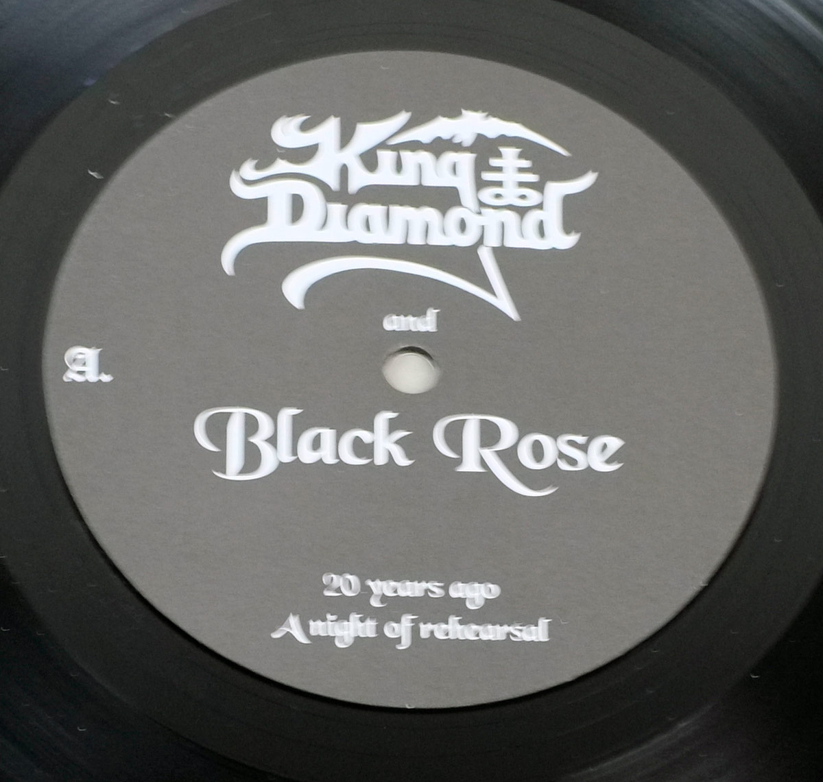 Close-up of Side One record label for King Diamond and Black Rose – 20 Years Ago – A Night of Rehearsal, showing a minimalist gray label design with the classic King Diamond logo in white gothic lettering centered at the top. Below it, the word 'and' appears in a smaller serif type, followed by the bold, sweeping title 'Black Rose' across the middle. At the bottom of the label, the album title '20 years ago – A night of rehearsal' is printed in delicate cursive script, completing the symmetrical composition. The letter 'A' marks the side designation on the left edge. The label’s simplicity, monochrome palette, and clean typography evoke the underground authenticity of limited-edition vinyl issues, emphasizing collectability and respect for analog purity.