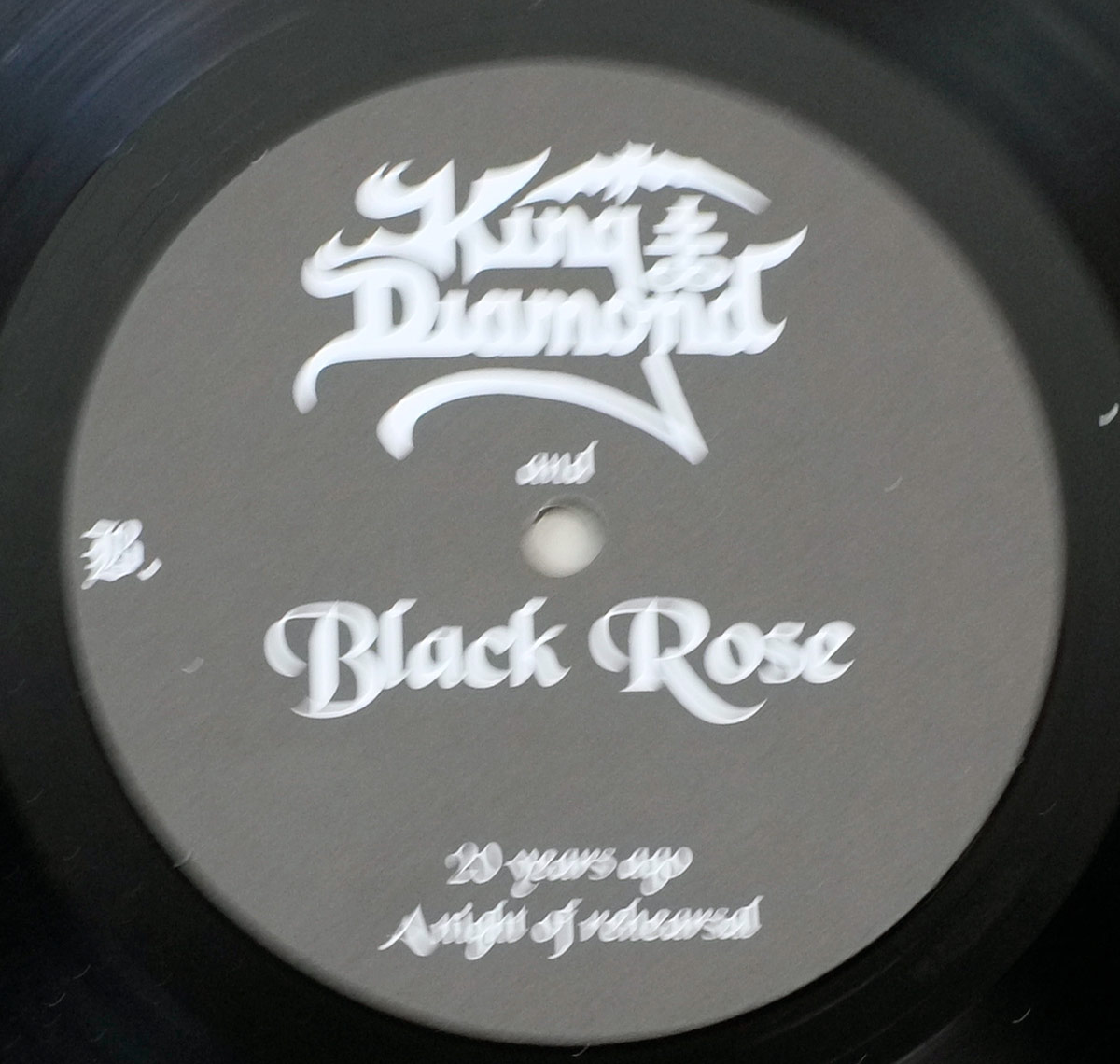Close-up of Side Two record label for King Diamond and Black Rose – 20 Years Ago – A Night of Rehearsal, displaying a matte gray surface with white gothic lettering. The King Diamond logo dominates the upper section, its stylized tail sweeping beneath the text. Centered below it, the smaller word 'and' connects to the band name 'Black Rose,' printed in ornate script. The album title '20 years ago – A night of rehearsal' appears in fine serif lettering near the bottom, balancing the circular layout. The letter 'B' on the left identifies the side. The clean, symmetrical design mirrors Side One’s label, maintaining visual continuity while emphasizing the collector aesthetic of this limited-edition Danish release.