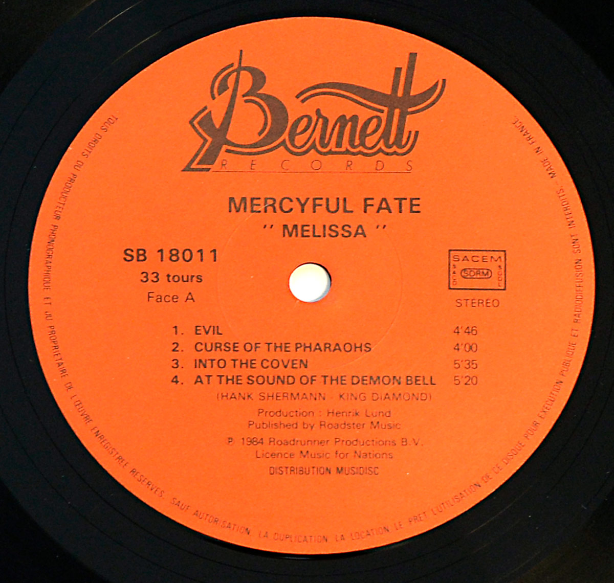 Close-up of the record label for Side A of Mercyful Fate’s 1983 album “Melissa,” from a French pressing by Bernett Records. The label is bright orange with black text. At the top, the Bernett Records logo is displayed in an elegant script. Below, “Mercyful Fate” and “Melissa” are printed in bold capital letters. The catalog number “SB 18011” is on the left, along with “33 tours” (33 RPM) and “Face A” (Side A). The tracklist includes “Evil,” “Curse of the Pharaohs,” “Into the Coven,” and “At the Sound of the Demon Bell,” with songwriting credits to Hank Shermann and King Diamond. Additional details include the “SACEM” logo, a “STEREO” marking, production credits for Henrik Lund, and a 1984 copyright notice for Roadrunner Productions B.V. The label also mentions “Licence Music for Nations” and “Distribution Musicdisc.