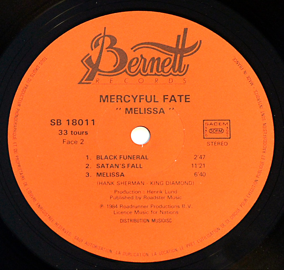 Close-up of the record label for Side B of Mercyful Fate’s 1983 album “Melissa,” from a French pressing by Bernett Records. The label is bright orange with black text. At the top, the Bernett Records logo is displayed in an elegant script. Below, “Mercyful Fate” and “Melissa” are printed in bold capital letters. The catalog number “SB 18011” is on the left, along with “33 tours” (33 RPM) and “Face 2” (Side 2). The tracklist includes “Black Funeral,” “Satan’s Fall,” and “Melissa,” with songwriting credits to Hank Shermann and King Diamond. Additional details include the “SACEM” logo, a “STEREO” marking, production credits for Henrik Lund, and a 1984 copyright notice for Roadrunner Productions B.V. The label also mentions “Licence Music for Nations” and “Distribution Musicdisc.”