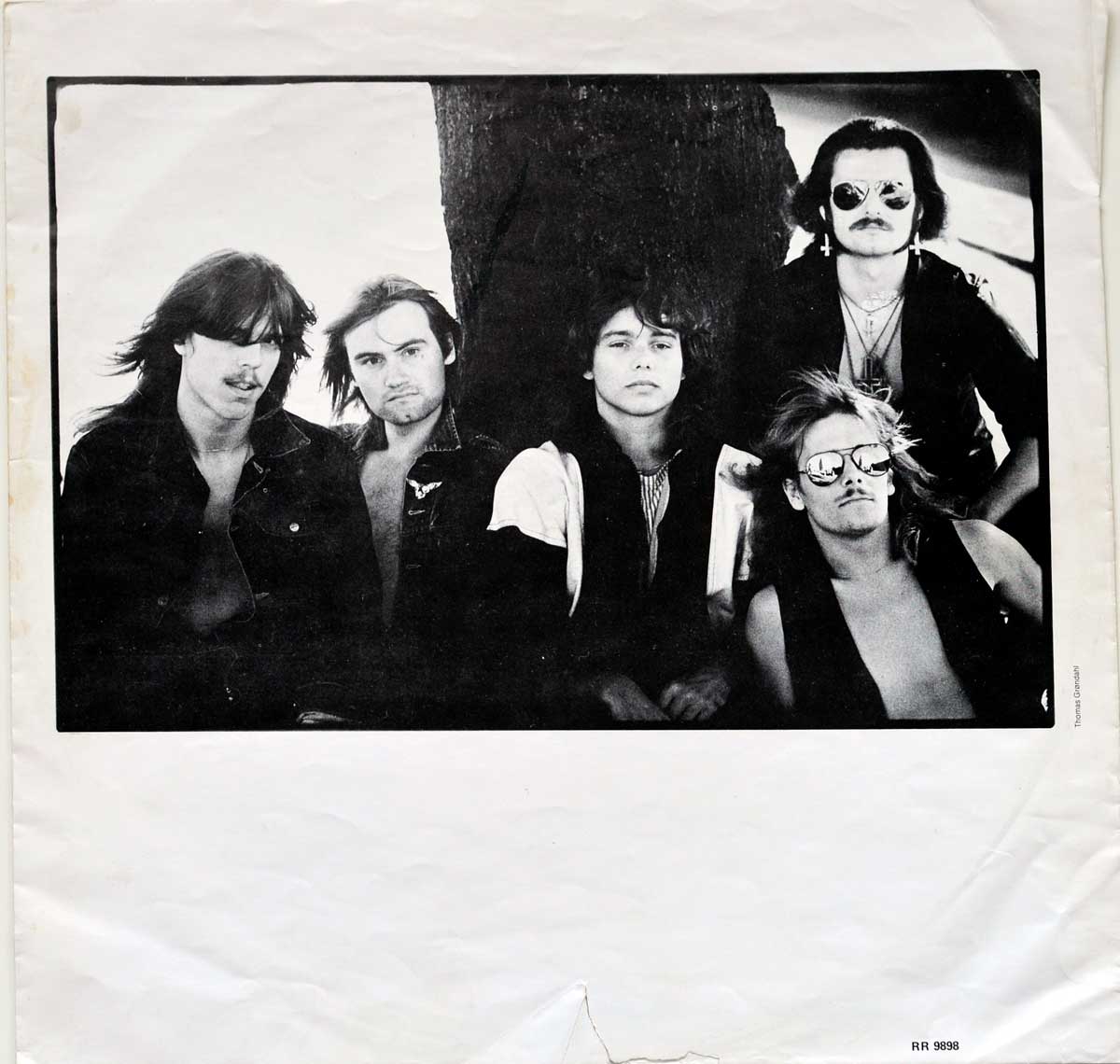 Black-and-white photograph on the custom inner sleeve of Mercyful Fate’s 1983 album Melissa. The image features the five band members posing outdoors against a large tree, exuding a dark and rebellious aura. From left to right: Kim Ruzz (drums) with long hair and an open jacket, Hank Shermann (lead guitar) with an intense gaze, Michael Denner (lead guitar) in a black-and-white jacket, King Diamond (vocals) wearing sunglasses, an inverted cross earring, and a leather jacket, and Timi Grabber (bass) shirtless under a vest with aviator sunglasses and wild hair. The high-contrast lighting enhances the moody and gothic feel of the image.  https://vinyl-records.nl