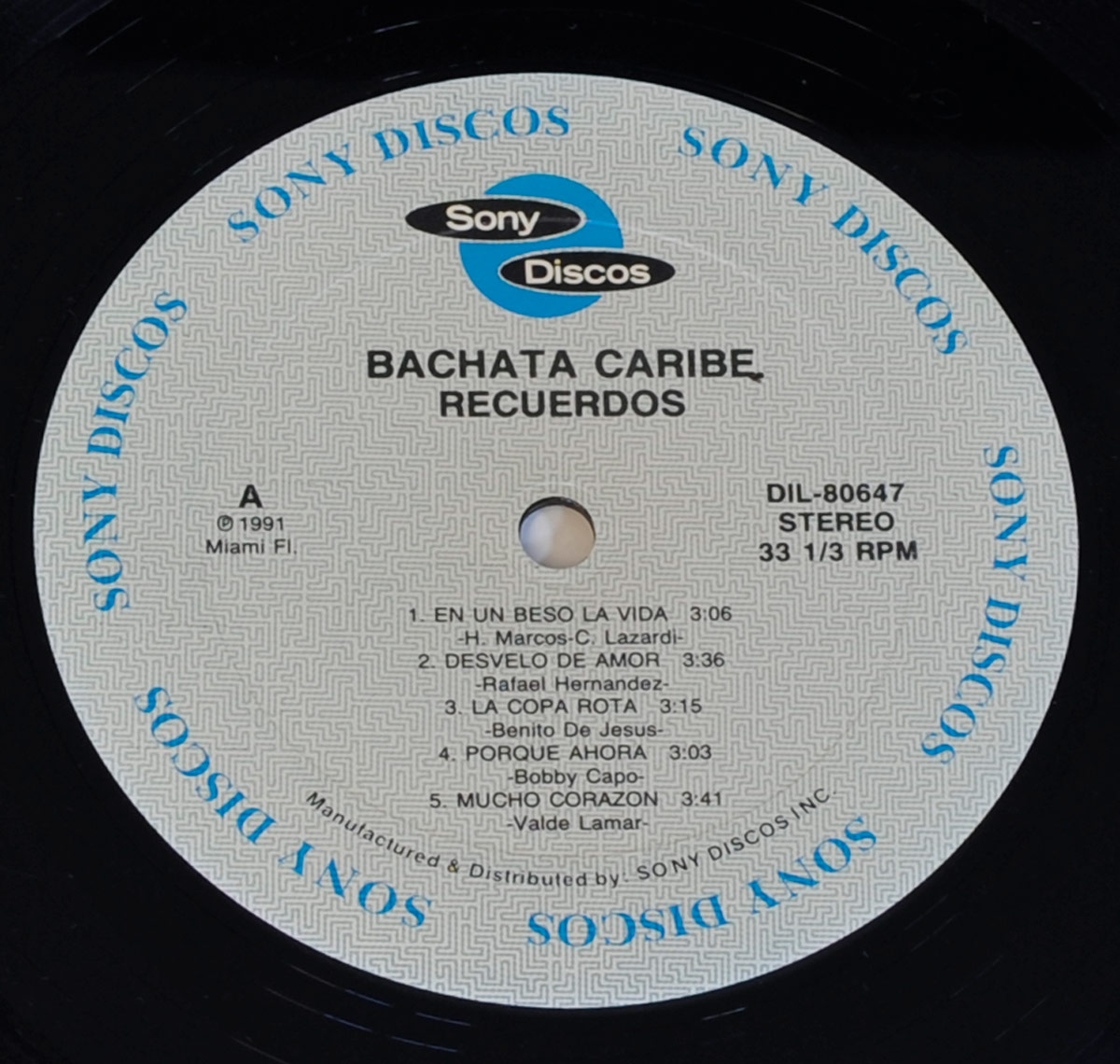 Close-up of the record label for the album 'Bachata Caribe - Recuerdos', released by Sony Discos. The label is circular with a light gray background featuring a subtle geometric pattern. The Sony Discos logo, with its black and blue design, is prominently displayed at the top center. The album title 'Bachata Caribe - Recuerdos' is printed in bold black capital letters beneath it. Around the edge of the label, 'SONY DISCOS' is printed multiple times in blue capital letters. The catalog number 'DIL-80647' and format 'STEREO 33 1/3 RPM' are positioned on the right side. The tracklist for Side A is neatly arranged in black text, listing songs such as 'En Un Beso La Vida', 'Desvelo de Amor', and 'La Copa Rota', each followed by the respective artist and duration. The bottom edge includes additional manufacturing and distribution details, indicating production by Sony Discos Inc. in Miami, Florida. The label's clean design ensures readability, balancing a professional aesthetic with the rhythmic essence of bachata music.