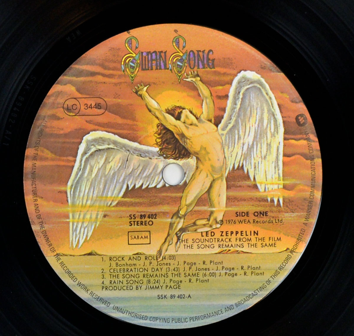 Close-up of the Side One label on a 12-inch vinyl record. The Swan Song artwork shows a winged human figure reaching upward against orange–gold sunset clouds. Ornate “Swan Song” lettering arcs at the top; a small LC 3445 mark appears in a circle to the left. Text blocks read SS 89 402, STEREO, SABAM; “Side One” and © 1976 WEA Records Ltd. Center text credits Led Zeppelin and the soundtrack title, lists four tracks with durations, and notes Produced by Jimmy Page. Matrix line at bottom: SSK 89 402-A. Perimeter carries standard legal text.