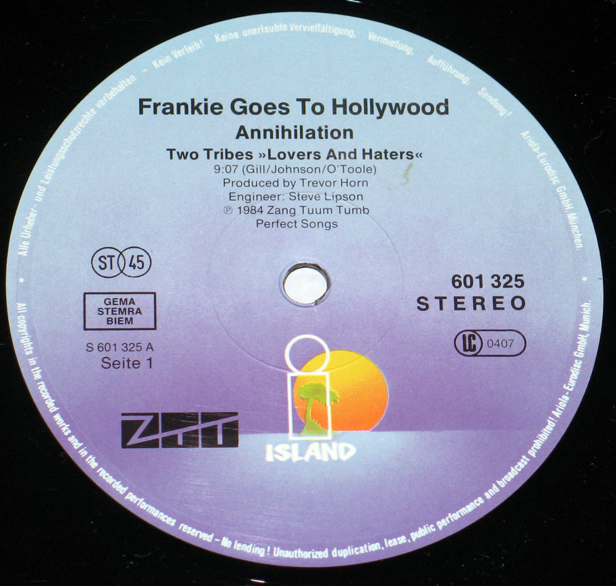 Close-up of Side One record label for Frankie Goes To Hollywood's Two Tribes 12 inch vinyl EP. The circular label is printed in a gradient of purple and light blue with the Island Records logo at the bottom: a stylized palm tree and orange sun within a white outline box. Text at the top states Frankie Goes To Hollywood Annihilation followed by the track title Two Tribes »Lovers And Haters« with credits to Gill, Johnson, and O'Toole. Production credits are also printed, listing Produced by Trevor Horn and Engineer: Steve Lipson. Copyright details include © 1984 Zang Tuum Tumb Perfect Songs. The left side contains the GEMA STEMRA BIEM rights society box, catalog number S 601 325 A, and notation Seite 1. The right side features the catalog number 601 325, Stereo designation, and LC 0407 label code. Around the edge of the label runs a white legal disclaimer prohibiting unauthorized duplication, lending, or broadcasting.