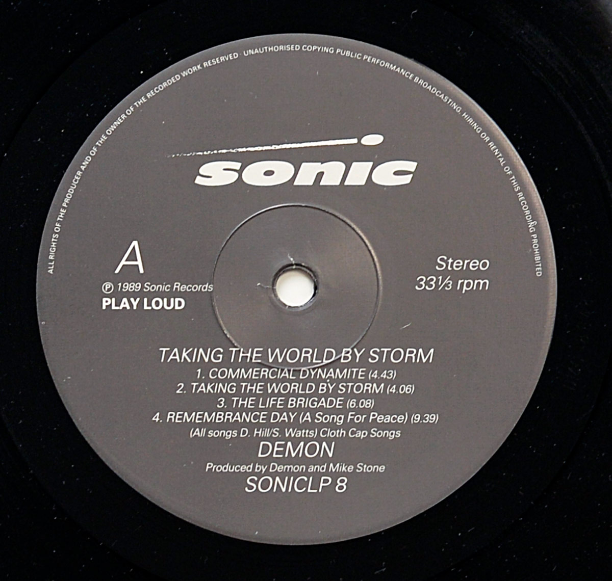 Close-up of Side One vinyl record label for Demon’s 1989 album Taking the World by Storm. The label is grey with white text, featuring the Sonic Records logo at the top. The words Stereo and 33⅓ rpm are printed on the right-hand side. The tracklist is displayed in the center, listing Commercial Dynamite (4:43), Taking the World by Storm (4:06), The Life Brigade (6:08), and Remembrance Day (A Song for Peace) (9:39). Beneath the tracklist are credits stating all songs were written by D. Hill and S. Watts, published by Cloth Cap Songs. The band name Demon is printed prominently, followed by production credits for Demon and Mike Stone. At the bottom is the catalog number SONICLP 8, along with a copyright notice for 1989 Sonic Records. The edge of the vinyl grooves encircles the label.