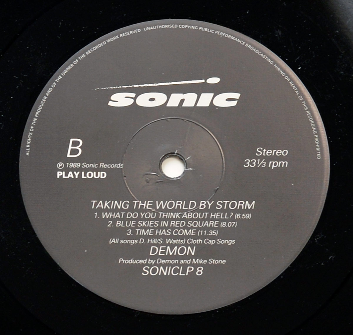Close-up of Side Two vinyl record label for Demon’s 1989 album Taking the World by Storm. The label is grey with white print, showing the Sonic Records logo at the top. The large letter B is positioned on the left-hand side, indicating this is Side Two. On the right, the words Stereo and 33⅓ rpm specify playback format. The tracklist includes What Do You Think About Hell? (6:59), Blue Skies in Red Square (8:07), and Time Has Come (11:35). Beneath the songs, credits confirm all tracks were written by D. Hill and S. Watts, published by Cloth Cap Songs. The band name Demon is centered in bold, followed by production credit to Demon and Mike Stone. The catalog number SONICLP 8 is printed at the bottom, along with a copyright notice © 1989 Sonic Records. The outer edge of the vinyl grooves surrounds the label.
