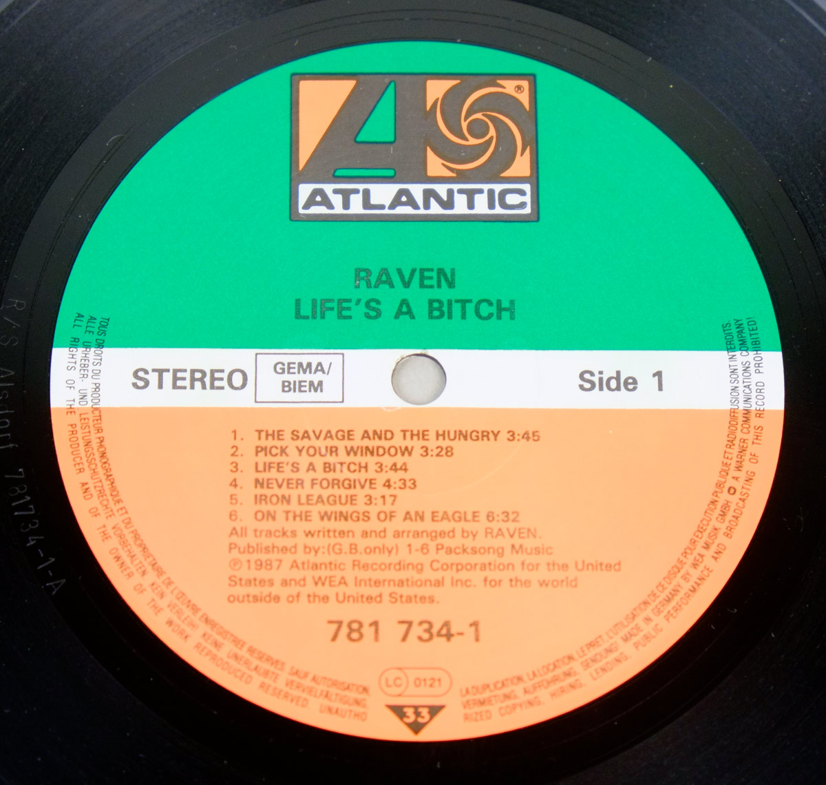 Close-up of Side One Atlantic record label for Raven - Life's A Bitch, showing the green, white, and orange Atlantic label on black vinyl. The label lists Raven, Life's A Bitch, Side 1, Stereo, GEMA/BIEM, catalog number 781 734-1, LC 0121, 33 RPM, six Side One tracks, copyright text, and Alsdorf matrix markings in the runout area.
