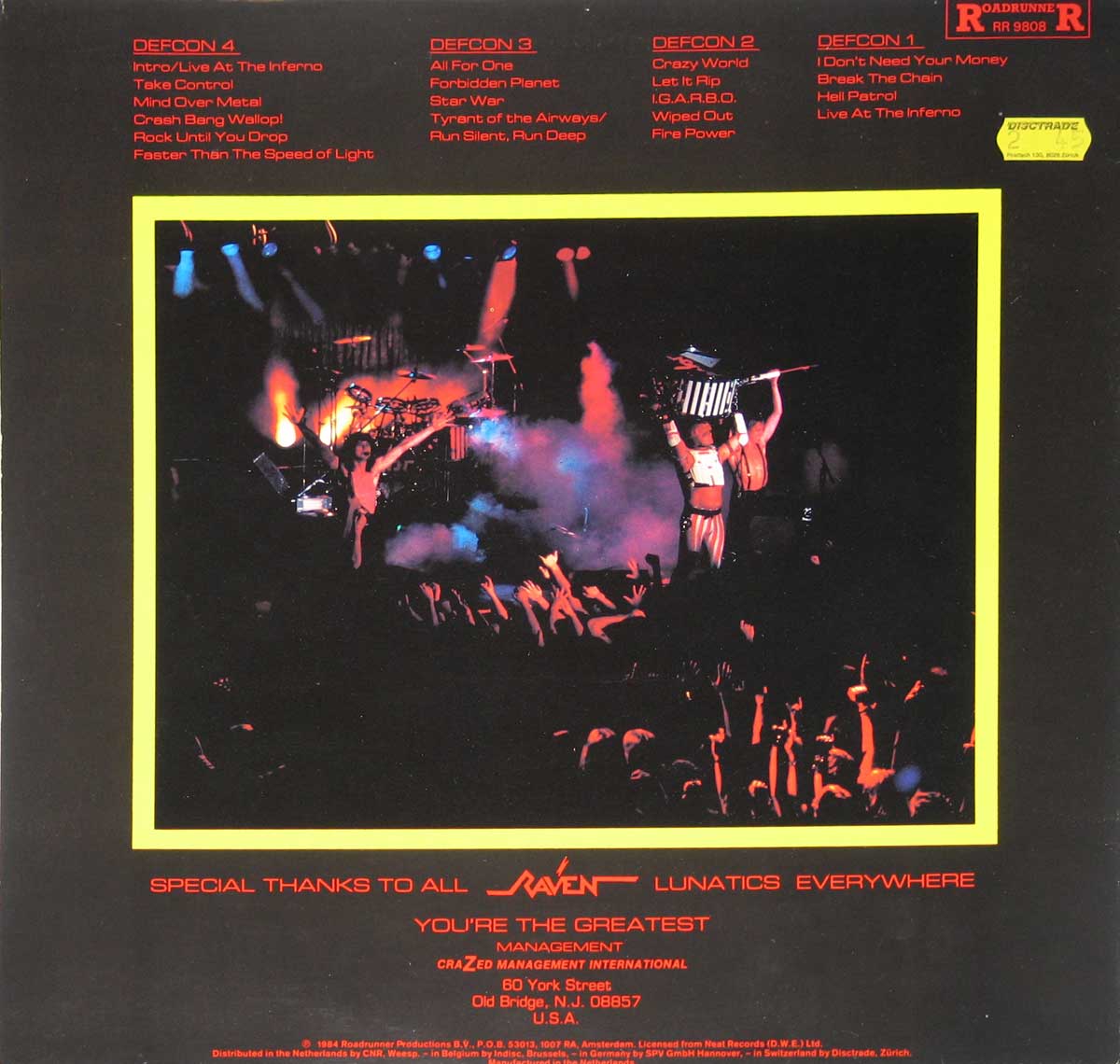 Back cover of Raven Live at the Inferno with red track listings across the top under DEFCON 4 to DEFCON 1 headings, a RoadrunneR RR 9808 logo at the upper right, and a yellow Distrade sticker nearby. A central live concert photo with a yellow border shows two shirtless musicians onstage with smoke, drums, lights, and raised crowd hands below.