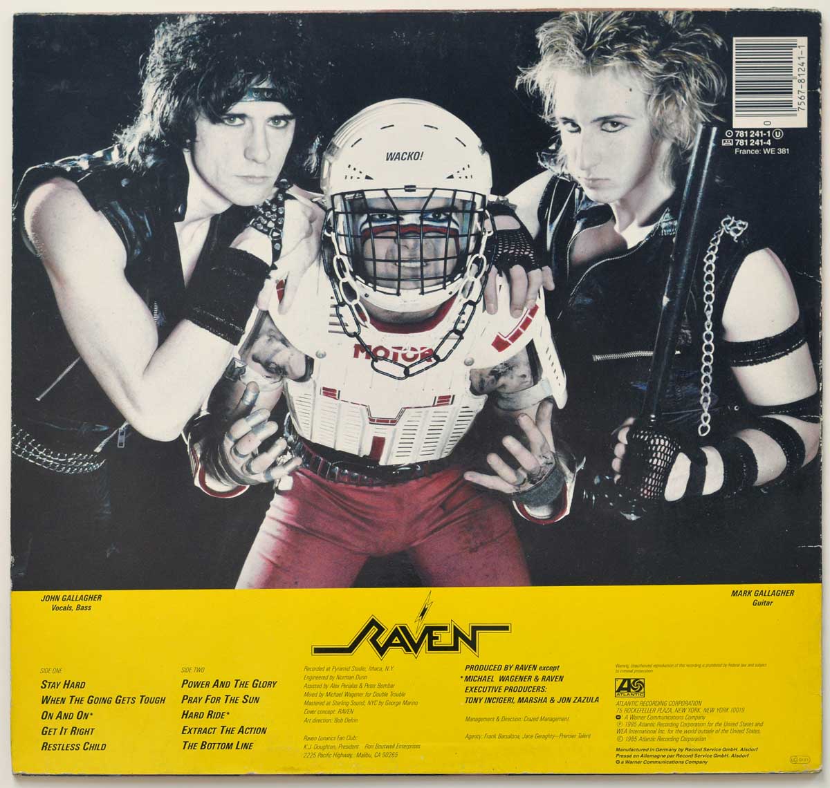 Raven Stay Hard back cover with John Gallagher on the left, Rob “Wacko” Hunter in hockey-style shoulder pads and helmet at centre, and Mark Gallagher on the right holding a bat. The upper image is a dark studio band photo, while the yellow lower panel carries the Raven logo, track columns, album credits, Atlantic Records logo, barcode, catalogue numbers, and German pressing text.