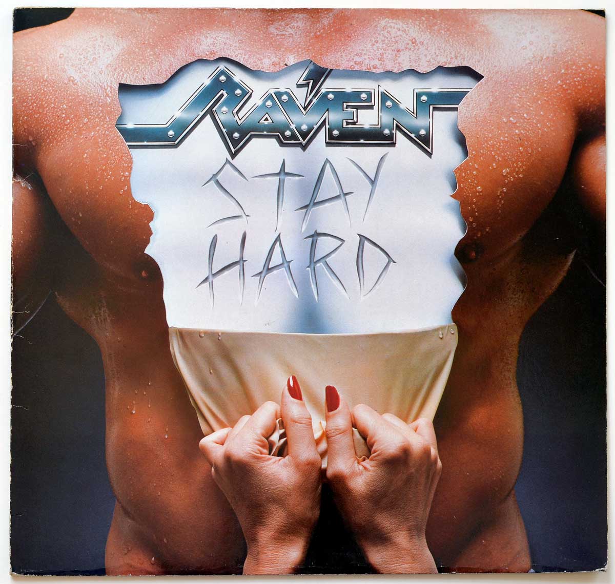 Album Front cover showing a close-cropped torso with water droplets across the skin, hands pulling down pale fabric, and a torn metallic panel across the chest carrying the chrome Raven logo and scratched Stay Hard title. The sleeve leans hard into mid-1980s shock-rock salesmanship: glossy, cheeky, sweaty, and about as subtle as a brick through a record-shop window.