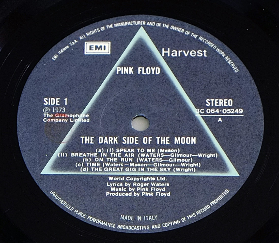 ‘Record label for an Italian pressing of Pink Floyd’s The Dark Side of the Moon, featuring a grey-blue background with a faint prism design. The label displays “Pink Floyd” and “The Dark Side of the Moon” at the center, with Side 1 track titles listed, including “Speak to Me,” “Breathe in the Air,” “On the Run,” “Time,” and “The Great Gig in the Sky.” The right side shows “Stereo” and catalogue number “3C 064-05249.” Copyright information and “Made in Italy” appear at the bottom.’