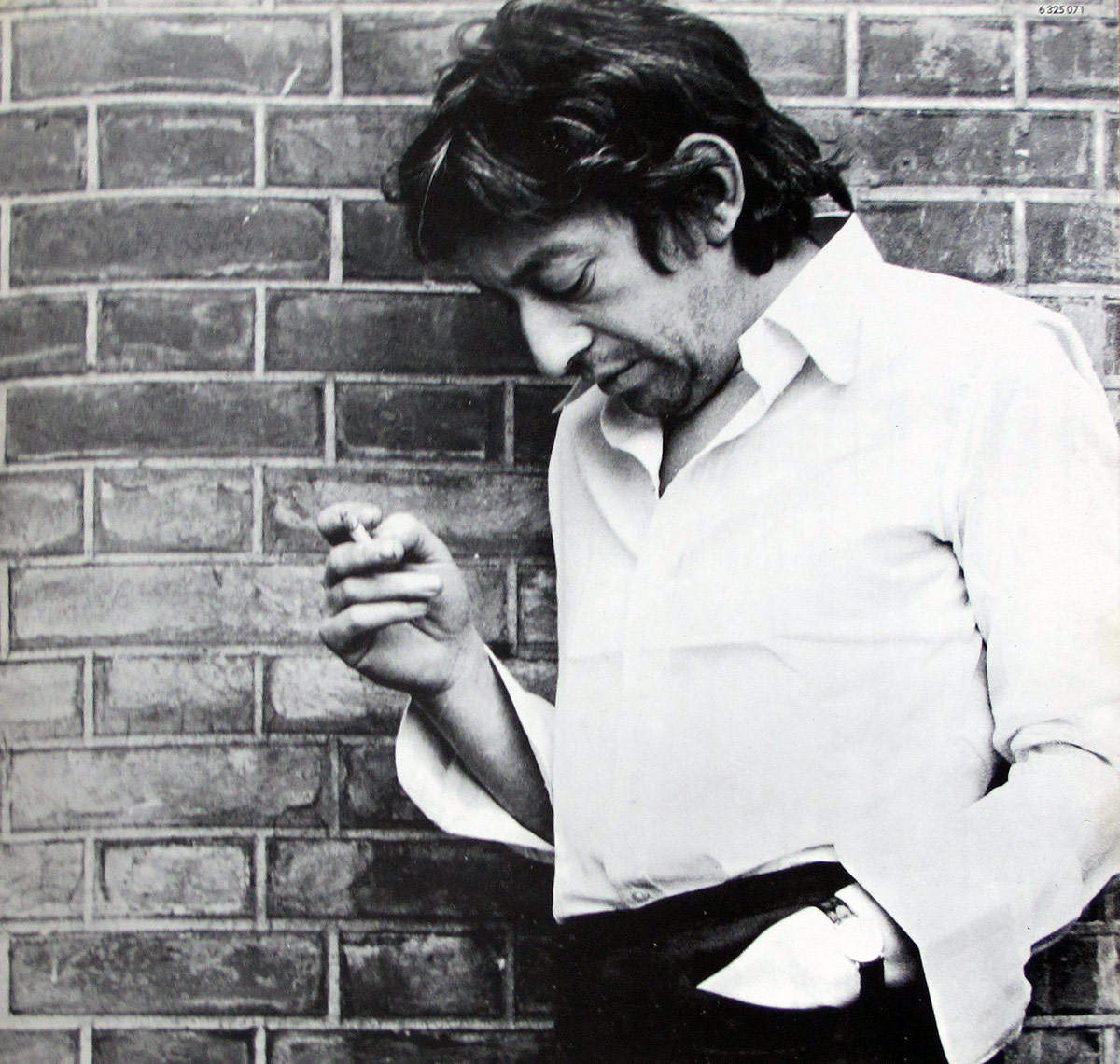 Inside gatefold photo from Serge Gainsbourg's 1971 LP showing Gainsbourg in profile, leaning against a brick wall, looking down while holding a cigarette in one hand, wearing an open-collared white shirt and dark trousers.