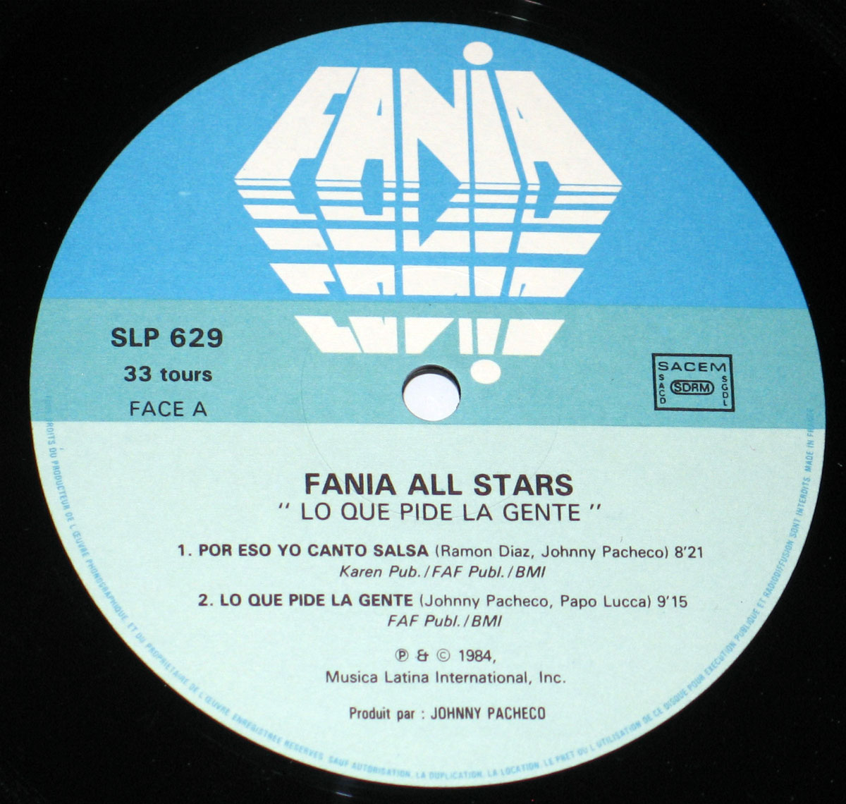 Close-up of the Side A record label for the Fania All Stars 1984 album Lo Que Pide La Gente. The circular label design features a light blue and white background with the bold geometric Fania logo across the top. The catalog number SLP 629 appears on the left, along with 33 tours and FACE A. The SACEM rights society logos are printed on the right. Centered below, the text reads Fania All Stars 'Lo Que Pide La Gente', listing two tracks: 'Por Eso Yo Canto Salsa' (Ramon Diaz, Johnny Pacheco) with an 8:21 duration, and 'Lo Que Pide La Gente' (Johnny Pacheco, Papo Lucca) with 9:15 duration. Publishing credits include Karen Pub., FAF Publ., and BMI. At the bottom, it notes ℗ & © 1984 Musica Latina International Inc. and Produced by Johnny Pacheco.