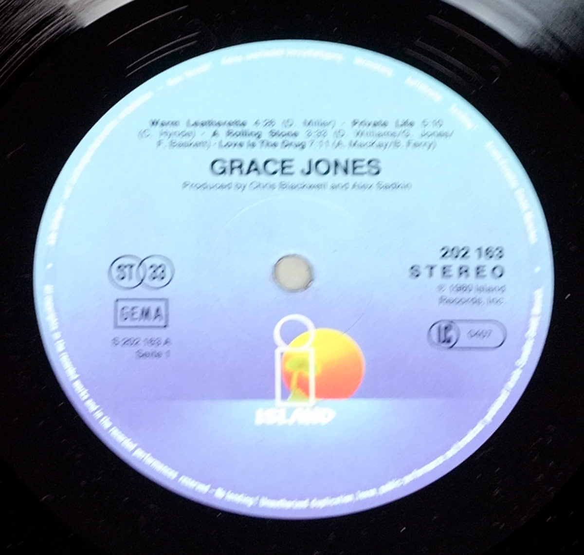 Close-up of the record label for Grace Jones’ 1980 album Warm Leatherette. The circular label features a gradient background fading from light blue at the top to a deeper violet at the bottom. Prominently at the lower center is the Island Records logo: a stylized palm tree within a rectangle, set against a vivid orange sun and yellow sky. At the top, the track listing for Side One is printed in small black text, including Warm Leatherette, Private Life, A Rolling Stone, and Love Is the Drug, along with songwriter credits and track durations. Centered below is the artist’s name GRACE JONES in bold uppercase letters, followed by the production credit Produced by Chris Blackwell and Alex Sadkin. On the right side, catalog number 202 163, the word STEREO, and the © 1980 Island Records, Inc. copyright notice are printed. On the left side are the GEMA rights society box, the stereo circle mark, and catalog matrix number S 202 163 A Serie 1. The label hole appears in the center, and the grooves of the black vinyl surround the printed area.