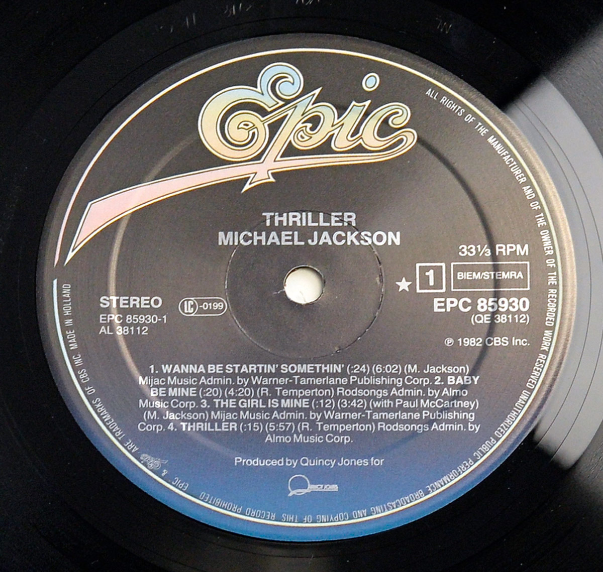 Close-up of the Side One record label from Michael Jackson’s Thriller vinyl LP, showing the bold Epic logo in yellow gradient lettering across the top. Below it, the text ‘Thriller – Michael Jackson’ appears in white capital letters centered above the track listing. Songs listed include ‘Wanna Be Startin’ Somethin’,’ ‘Baby Be Mine,’ ‘The Girl Is Mine’ (with Paul McCartney), and ‘Thriller.’ Additional details include the catalog number EPC 85930, BIEM/STEMRA rights designation, and ‘Made in Holland’ text printed along the rim. The background fades from dark gray to deep blue, characteristic of 1980s European Epic pressings, printed with crisp clarity and perfect alignment around the spindle hole.
