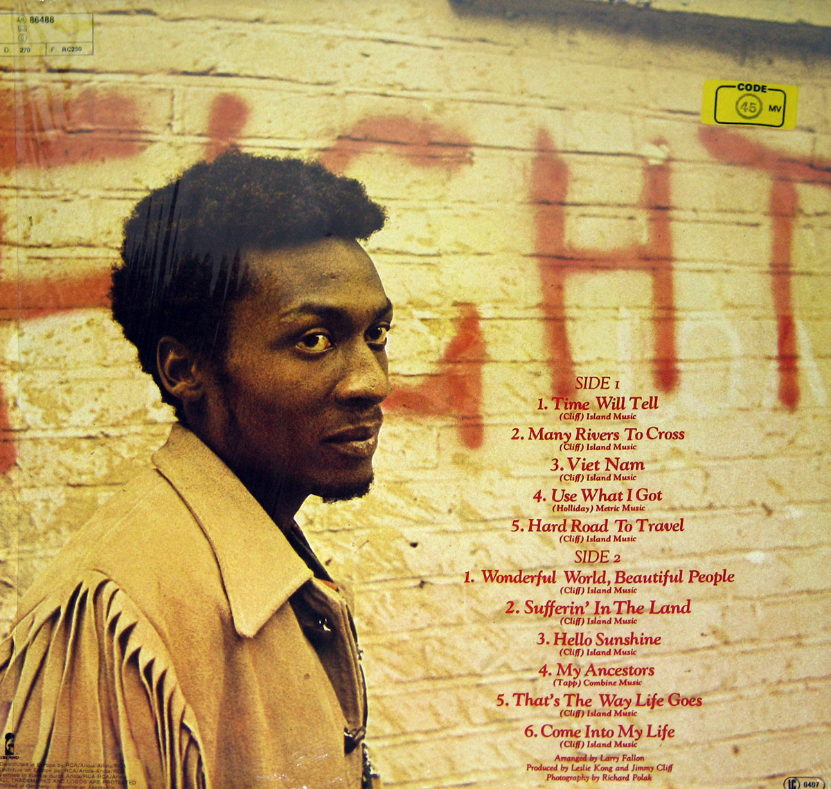 Back cover of the album Wonderful World Beautiful People by Jimmy Cliff. The photograph shows Cliff in profile, turning his head slightly towards the camera with an intent and watchful gaze. He wears a tan suede jacket with long fringe details along the sleeves and shoulders, standing against a cream-colored brick wall marked with red spray-painted graffiti. To the right of the image, the complete track listing for both sides of the record is printed in red text, numbered and titled clearly. Small production credits are listed at the bottom left, including arrangement by Larry Fallon, production by Leslie Kong and Jimmy Cliff, and photography by Richard Polak. Stickers for catalog number and code labels appear in the upper corners, giving the cover an authentic vintage vinyl appearance.