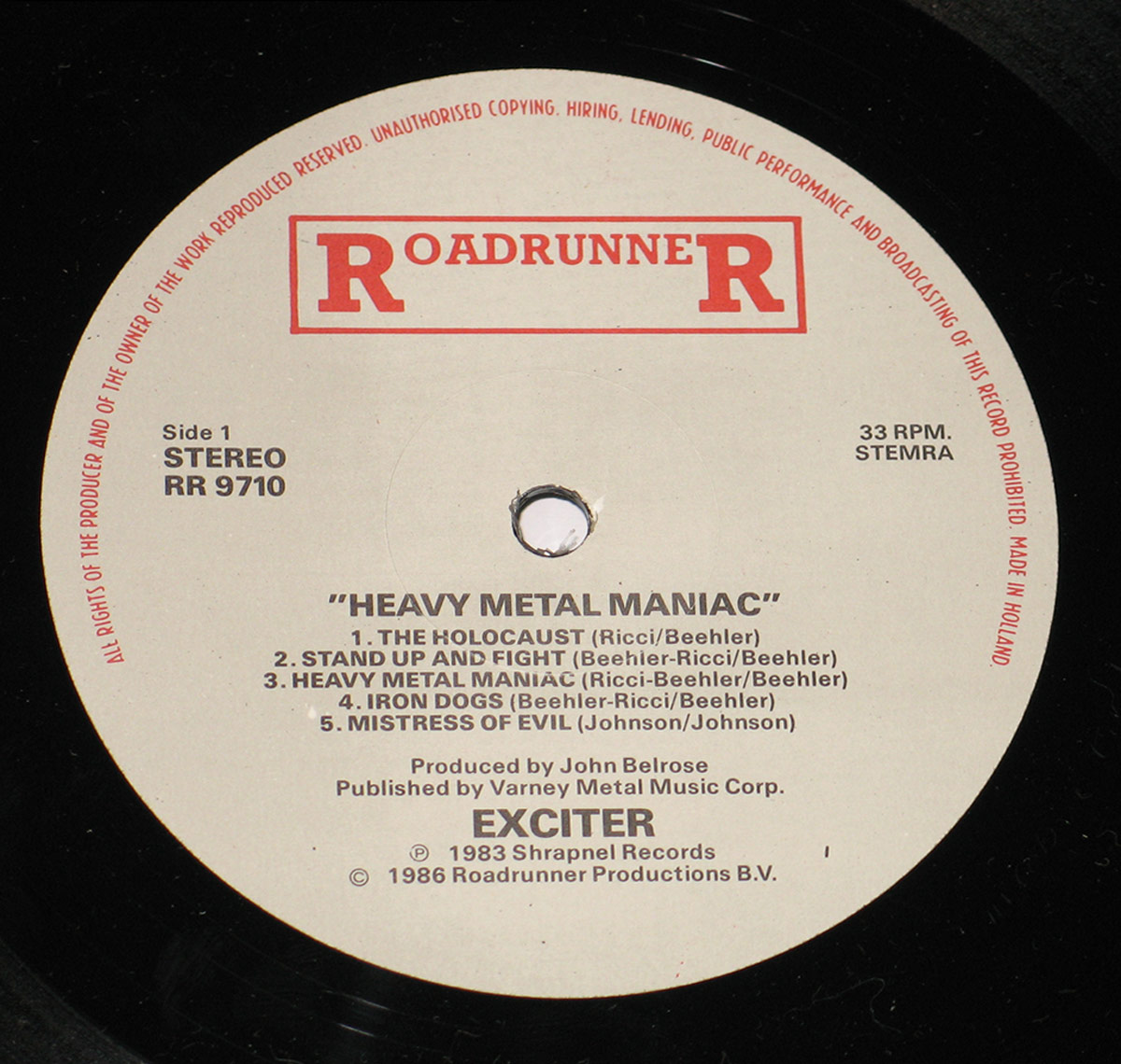 Side 1 label of Exciter's Heavy Metal Maniac vinyl record, released by Roadrunner Records. The off-white label features the bold red Roadrunner logo at the top, with track titles and credits printed in black, surrounding the center spindle hole. The catalog number RR 9710 and production details are also included.