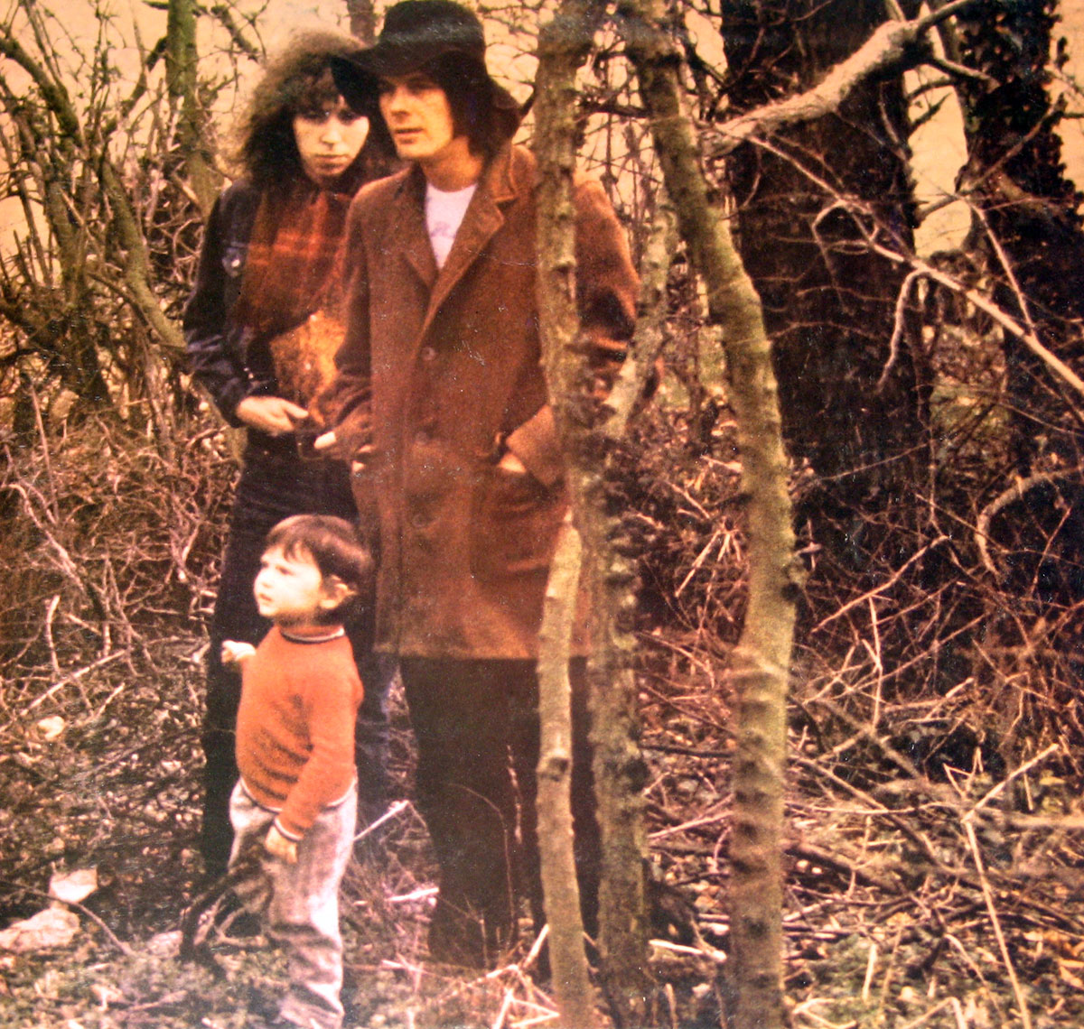 High resolution photo from the inside gatefold of Fairport Convention's 1970 album Full House. Two men and a small child stand among bare trees and tangled undergrowth. One man with long curly hair wears a dark jacket with a red-brown scarf, his hands in his pockets. Beside him, another man in a brown overcoat and wide-brimmed black hat looks outward, hands also in his pockets. In front, a young boy in a red sweater and grey trousers gazes upward, holding a stick. The woodland setting is raw and natural, with earthy muted tones.