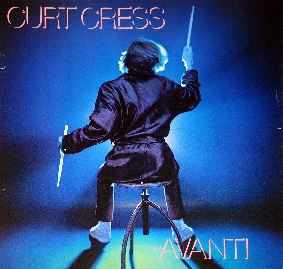 The album cover of Curt Cress's Avanti shows the drummer from behind, seated on a drum stool, illuminated in vibrant blue light. He wears a satin-like robe and holds drumsticks raised in both hands, as if in mid-performance. The scene is lit dramatically, casting strong shadows on the floor, creating a dynamic and theatrical atmosphere. The name CURT CRESS is positioned in large, stylized pink-red letters at the top left, and the album title AVANTI appears at the bottom right in matching style. The overall composition emphasizes motion, rhythm, and artistic flair. https://vinyl-records.nl