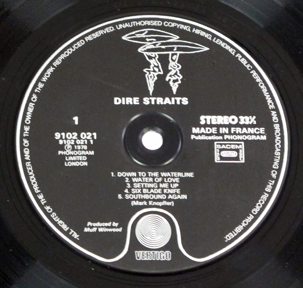 Close-up of the Side One label from the DIRE STRAITS – Self-Titled France Black Vertigo 2nd Pressing. The label is matte black with bold white print, dominated at the top by Vertigo’s iconic ‘two-mushroom/UFO’ logo: two rounded, flying-saucer shapes stacked with jagged lightning-bolt stalks extending downward. This graphic was used as an alternative to the swirl logo on specific late-1970s French Vertigo releases. The band name appears in uppercase directly beneath the artwork, centered above the tracklist. To the left are the catalogue number 9102 021, the year © 1978, and the PHONOGRAM LIMITED LONDON imprint. To the right are the STEREO 33⅓ speed marking, ‘MADE IN FRANCE’, and SACEM rights box. The bottom features the classic Vertigo swirl emblem within a curved frame. Rim text encircles the label, warning against copying, hiring or public performance without permission.