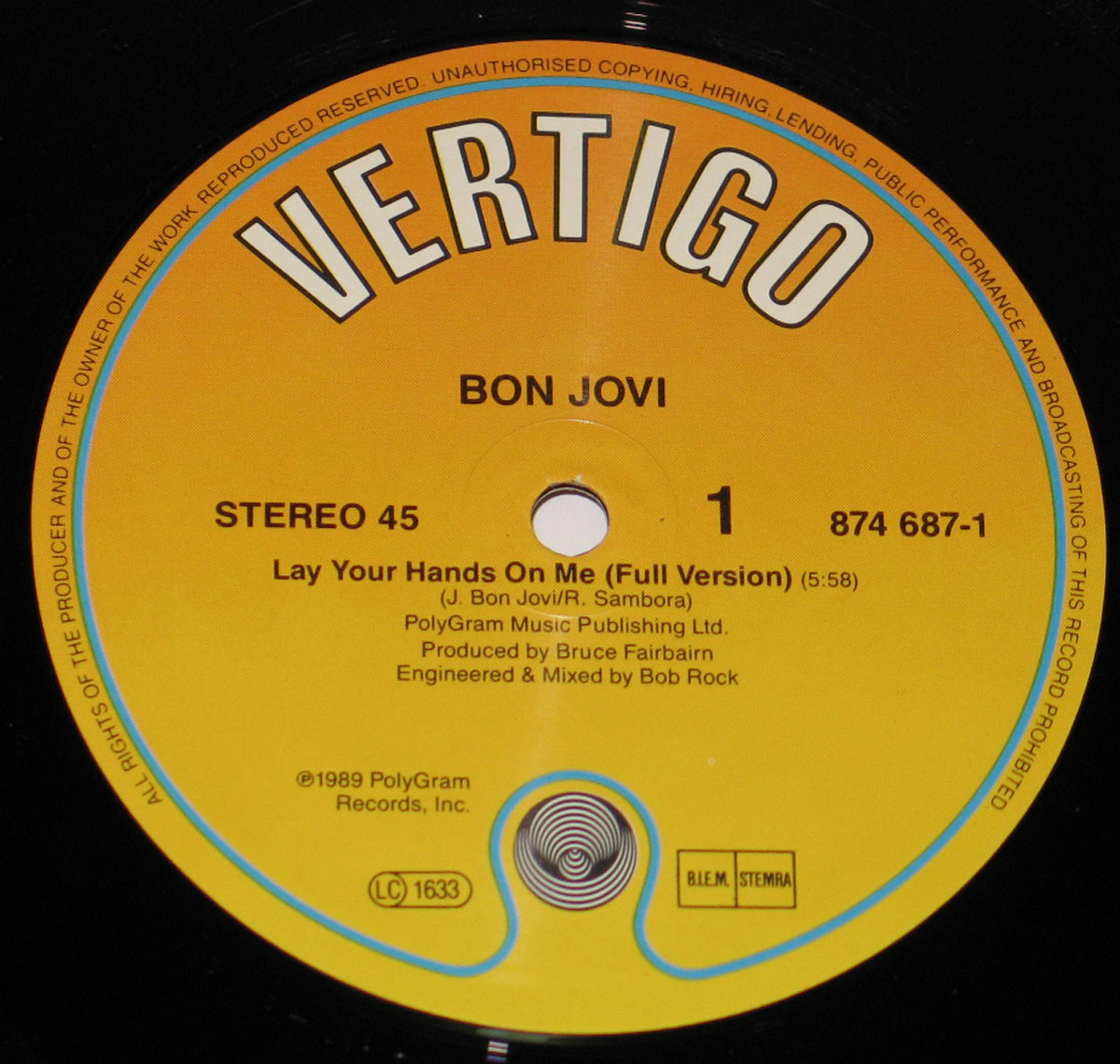 Side One record label of Bon Jovi’s 1989 12-inch vinyl EP Lay Your Hands On Me. The label is printed in a vivid orange and yellow gradient, characteristic of Vertigo Records releases of the era. Across the top, the large bold word VERTIGO curves along the edge, immediately recognizable as the label’s iconic branding. Below, the band’s name BON JOVI is printed in uppercase letters, followed by the track title Lay Your Hands On Me (Full Version) with a timing of 5:58. Songwriting credits list J. Bon Jovi and R. Sambora, with publishing under PolyGram Music Publishing Ltd. Additional lines give production credit to Bruce Fairbairn and mixing/engineering credit to Bob Rock. The catalog number 874 687-1 appears on the right-hand side, with legal copyright details around the circumference, marking this as an authentic 1989 PolyGram pressing.