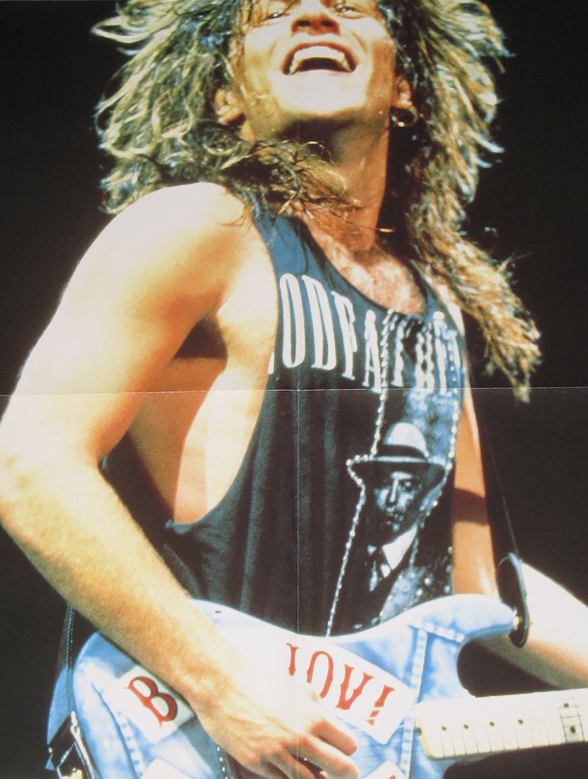Fold-out poster included with Bon Jovi’s 1989 12-inch vinyl EP Lay Your Hands On Me. The image shows Jon Bon Jovi mid-performance, smiling joyfully with his head tilted back and long blond hair flowing wildly. He wears a sleeveless black tank top with a bold print featuring an old-fashioned gangster in a suit and fedora. Slung across his body is a custom light blue electric guitar decorated with the band’s name BON JOVI in large red letters across its surface. The shot is tightly cropped on his upper body, conveying motion, sweat, and the exhilaration of a live arena performance during the peak of the band’s late-1980s fame.
