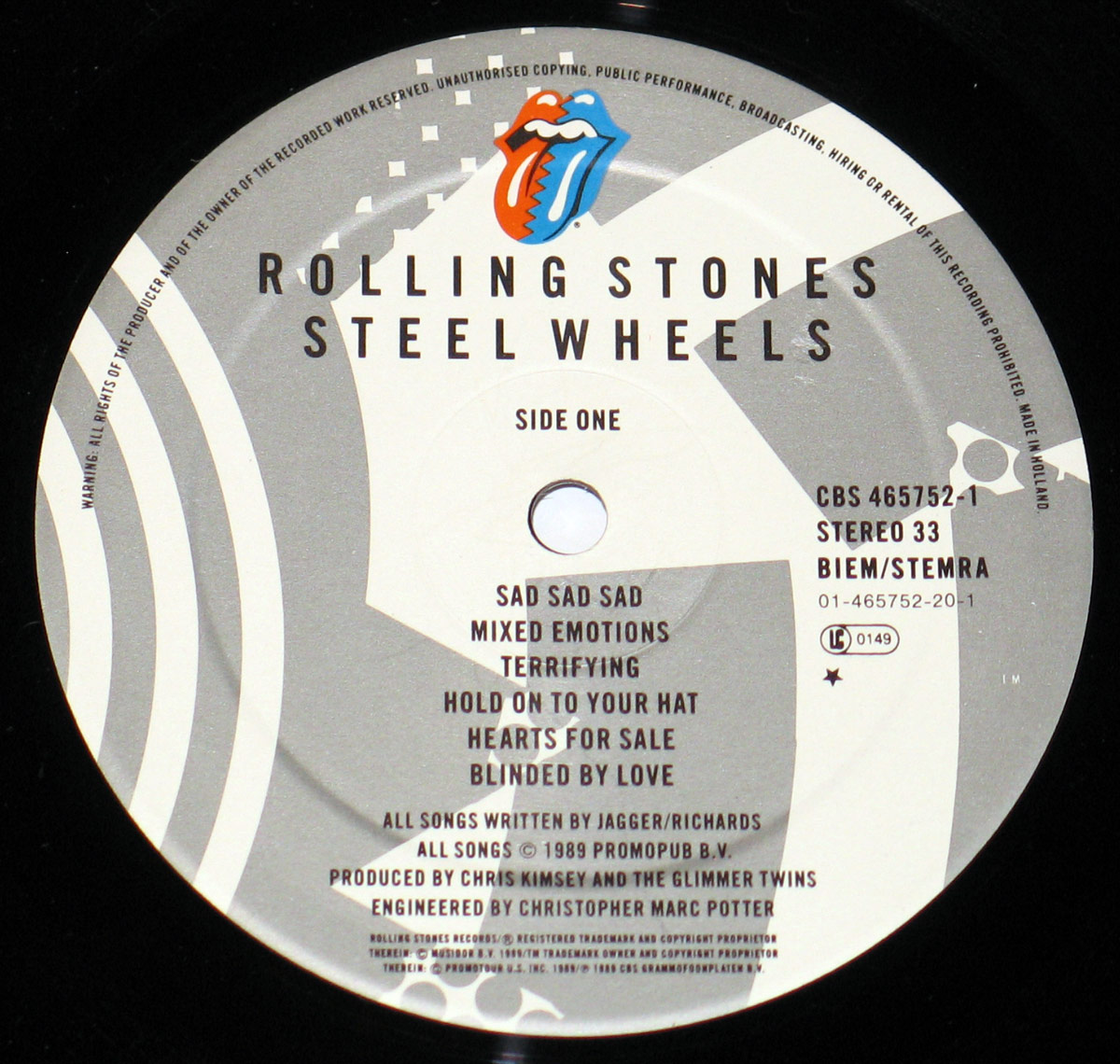 Close-up of the Side One record label of the Rolling Stones album Steel Wheels. The label is printed in shades of gray with angular graphic patterns, featuring the iconic red tongue and lips logo at the top. The text Rolling Stones Steel Wheels is prominently displayed in black uppercase letters, centered near the top. Below it, Side One is indicated, followed by the full track listing: Sad Sad Sad, Mixed Emotions, Terrifying, Hold On to Your Hat, Hearts for Sale, and Blinded by Love. Production credits, catalog information, and copyright details are printed around the edges, including CBS 465752-1, Stereo 33, BIEM/STEMRA, and the phrase Made in Holland.