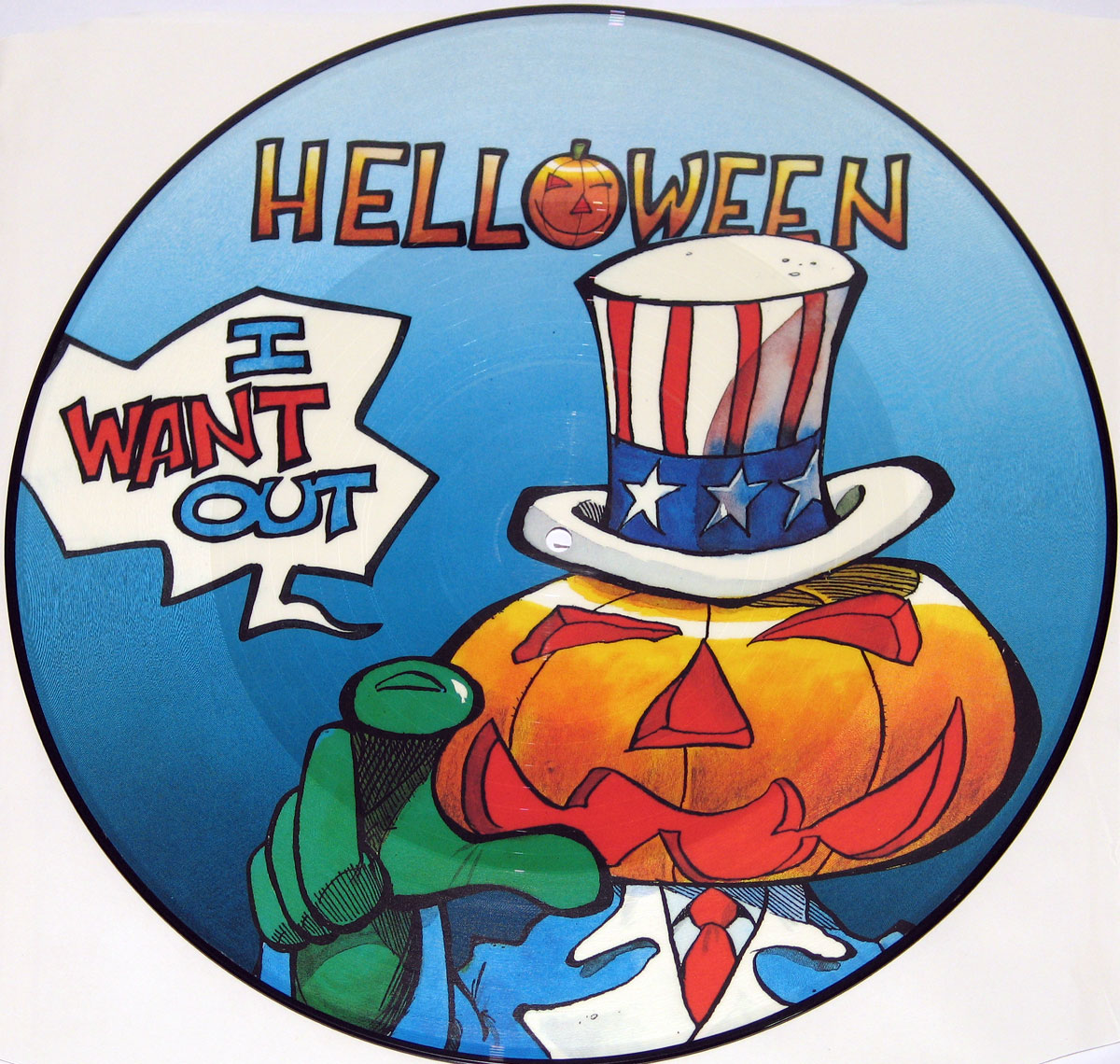 A round picture disc featuring Helloween’s I Want Out single. The artwork depicts a cartoonish jack-o’-lantern-headed figure dressed as Uncle Sam, wearing a red, white, and blue top hat and suit. The character points directly at the viewer with a green-gloved hand. A speech bubble in bold comic-style font reads “I WANT OUT.” The background is bright blue, and the band’s name, Helloween, appears at the top in a playful, slightly distorted font, with the “O” replaced by a carved pumpkin face.